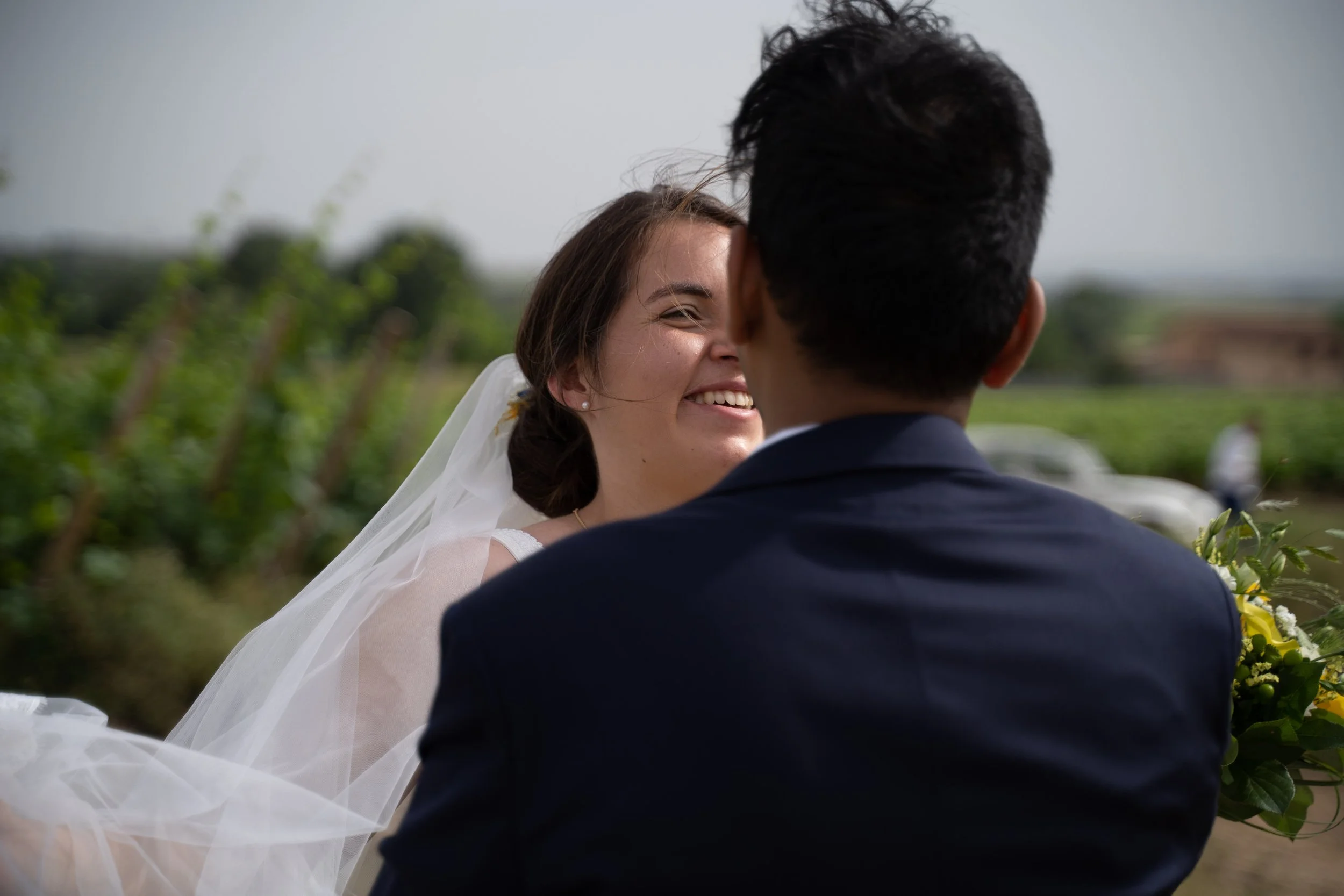 Un couple marié partage un moment dans une nature verdoyante, la femme sourit tendrement, vêtue d'une robe de mariage blanche et d'un voile, tandis que l'homme, de dos, porte un costume foncé.