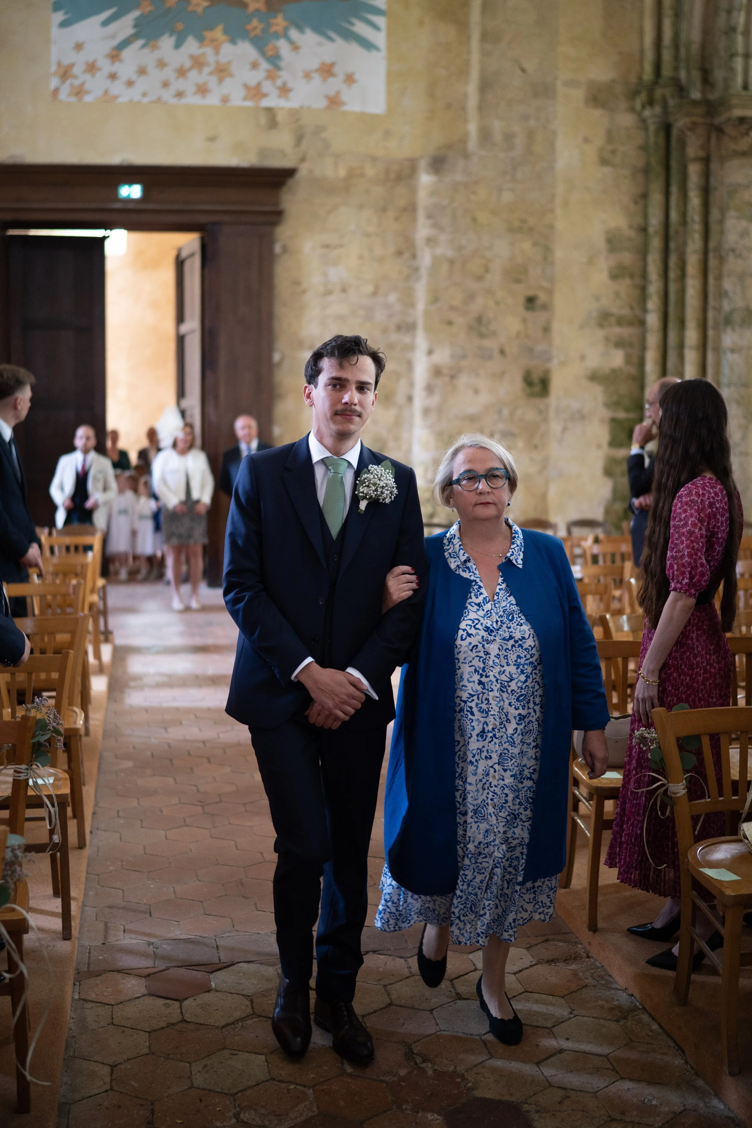 Un jeune homme en costume avec une boutonnière de fleurs, accompagnée d'une femme âgée portant des lunettes et une robe bleue avec un manteau coordonné, marchant dans une église lors d'une cérémonie, probablement un mariage.