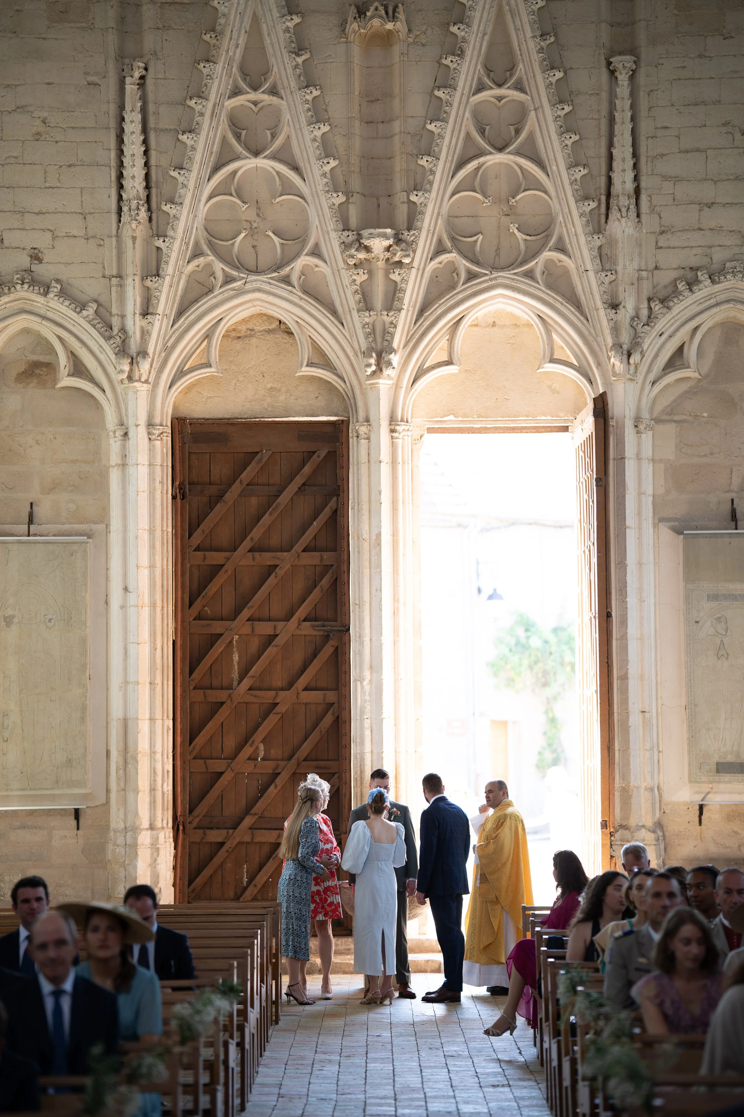 Une cérémonie de mariage dans une église avec des invités assis sur des bancs en bois et un groupe de personnes debout près de la porte en haut, sous une architecture gothique avec des détails sculptés et des vitraux.