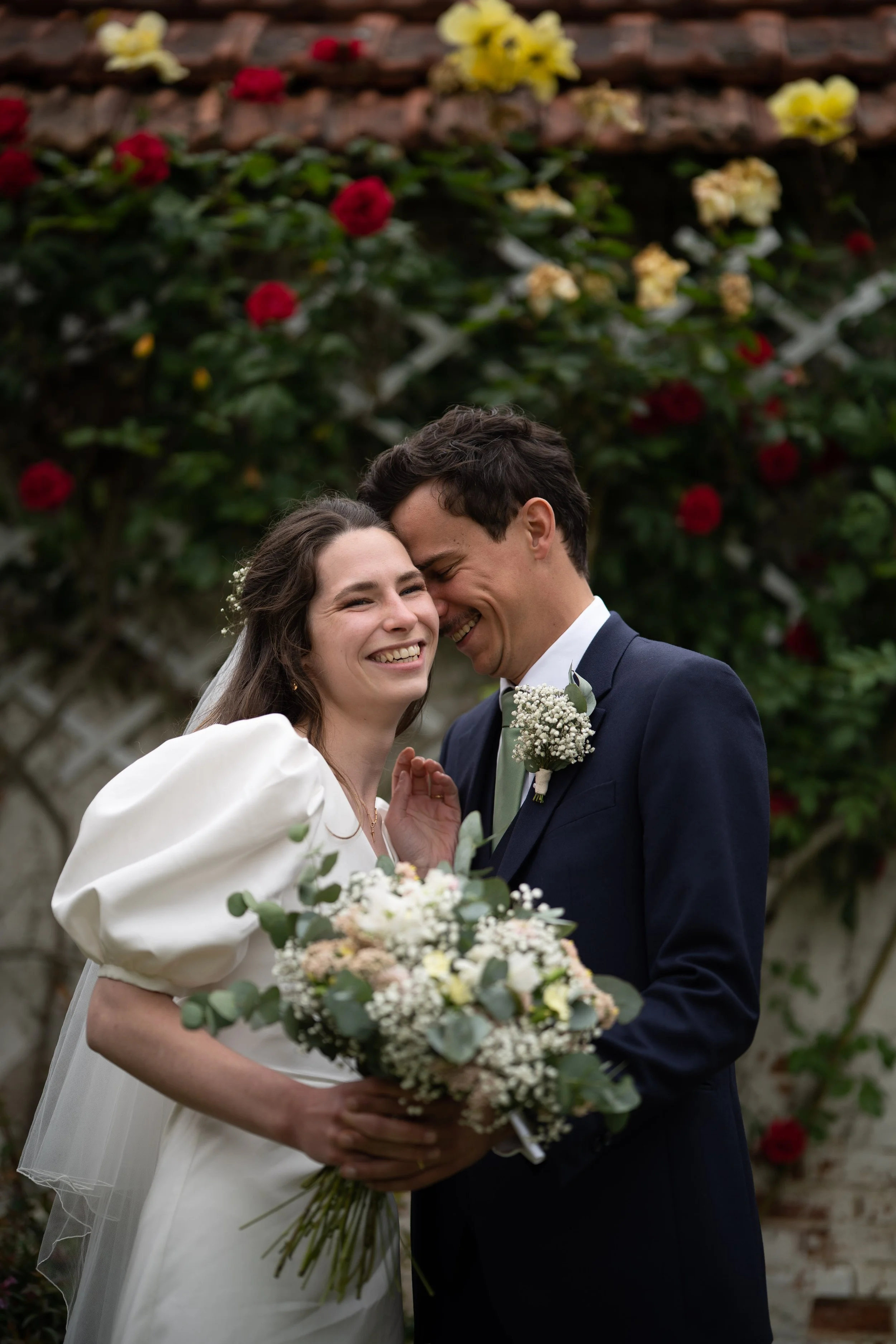 Un couple de mariés souriant lors de leur mariage, la femme en robe blanche tenant un bouquet de fleurs et l'homme en costume noir avec une boutonnière, devant un mur recouvert de fleurs rouges, jaunes et blanches.