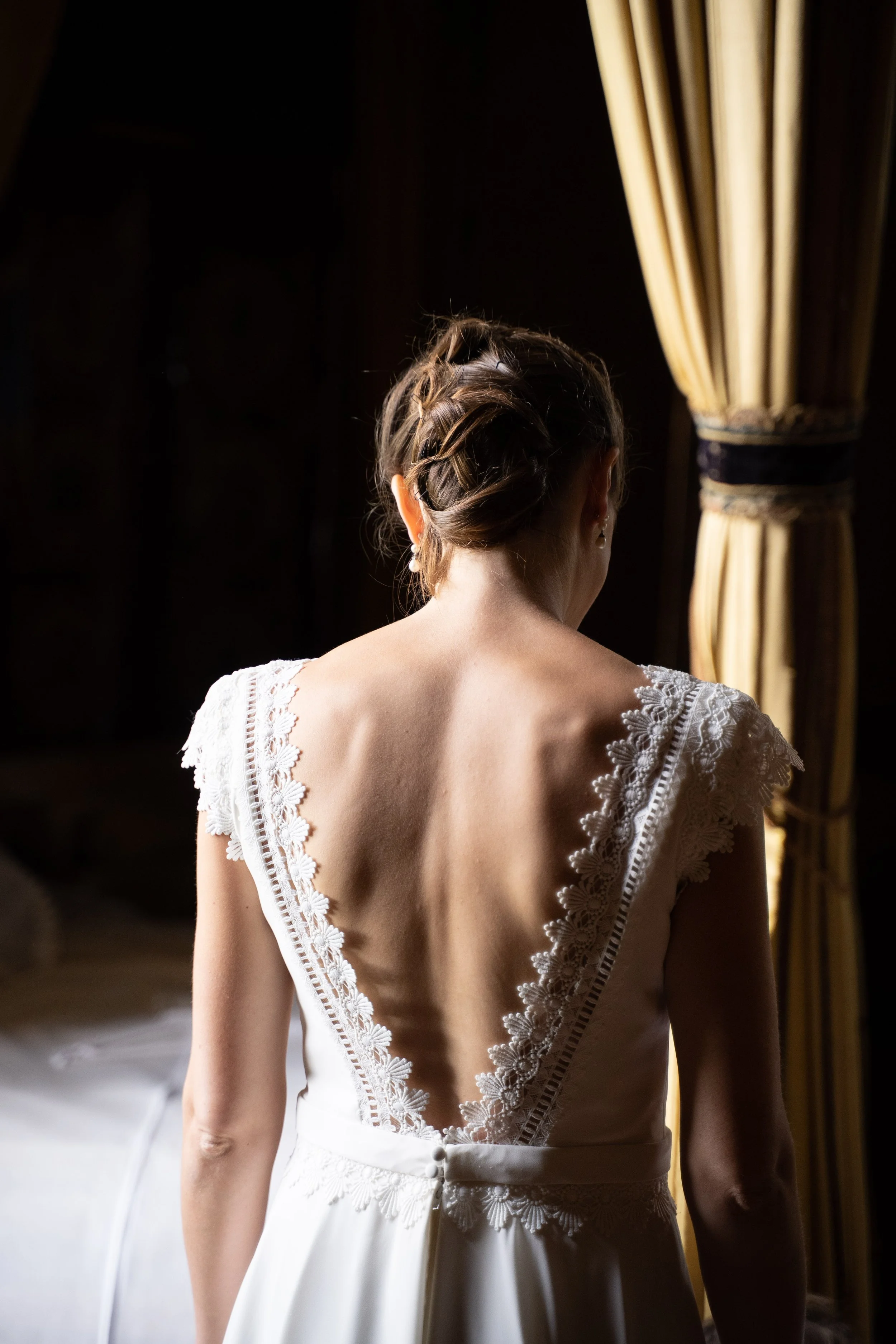 Une femme vue de dos dans une robe blanche en dentelle, près de rideaux dorés, dans une pièce sombre.