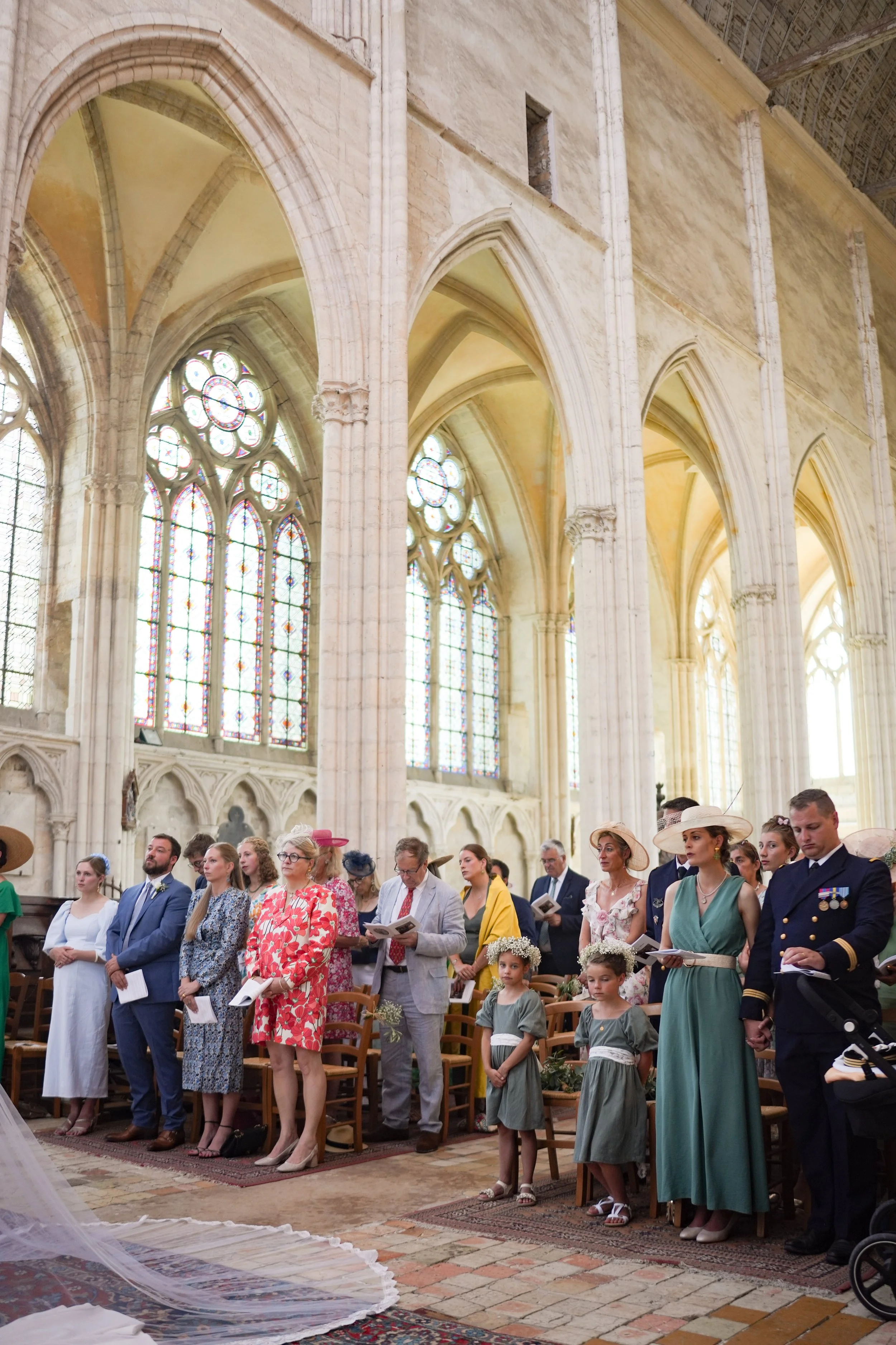 Groupe de personnes lors d'une cérémonie religieuse dans une église gothique, avec vitraux colorés, murs en pierre et arches hautes, certains portant des vêtements élégants et des chapeaux.