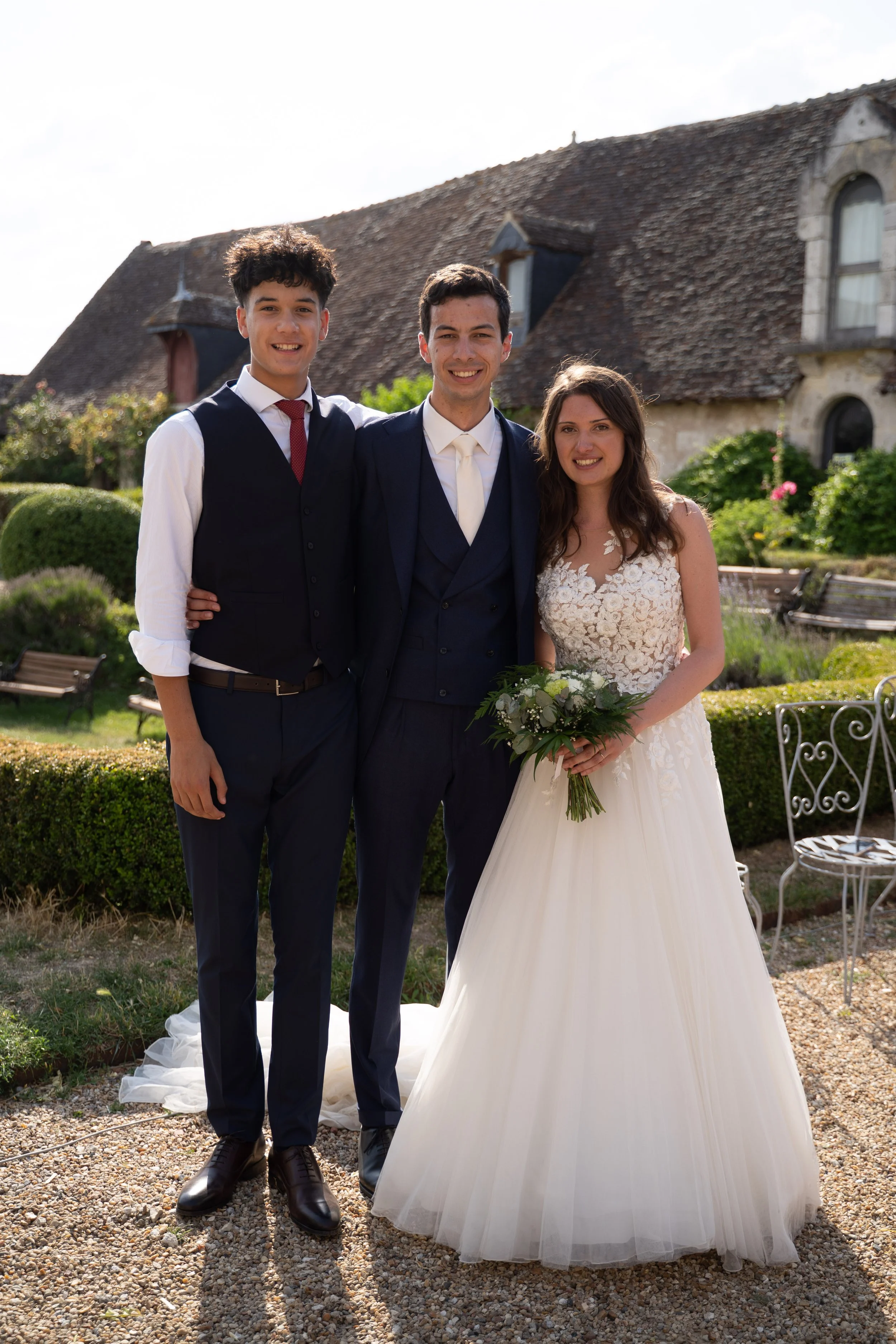 Trois jeunes personnes en tenue de mariage posant dans un jardin devant une maison en pierre.