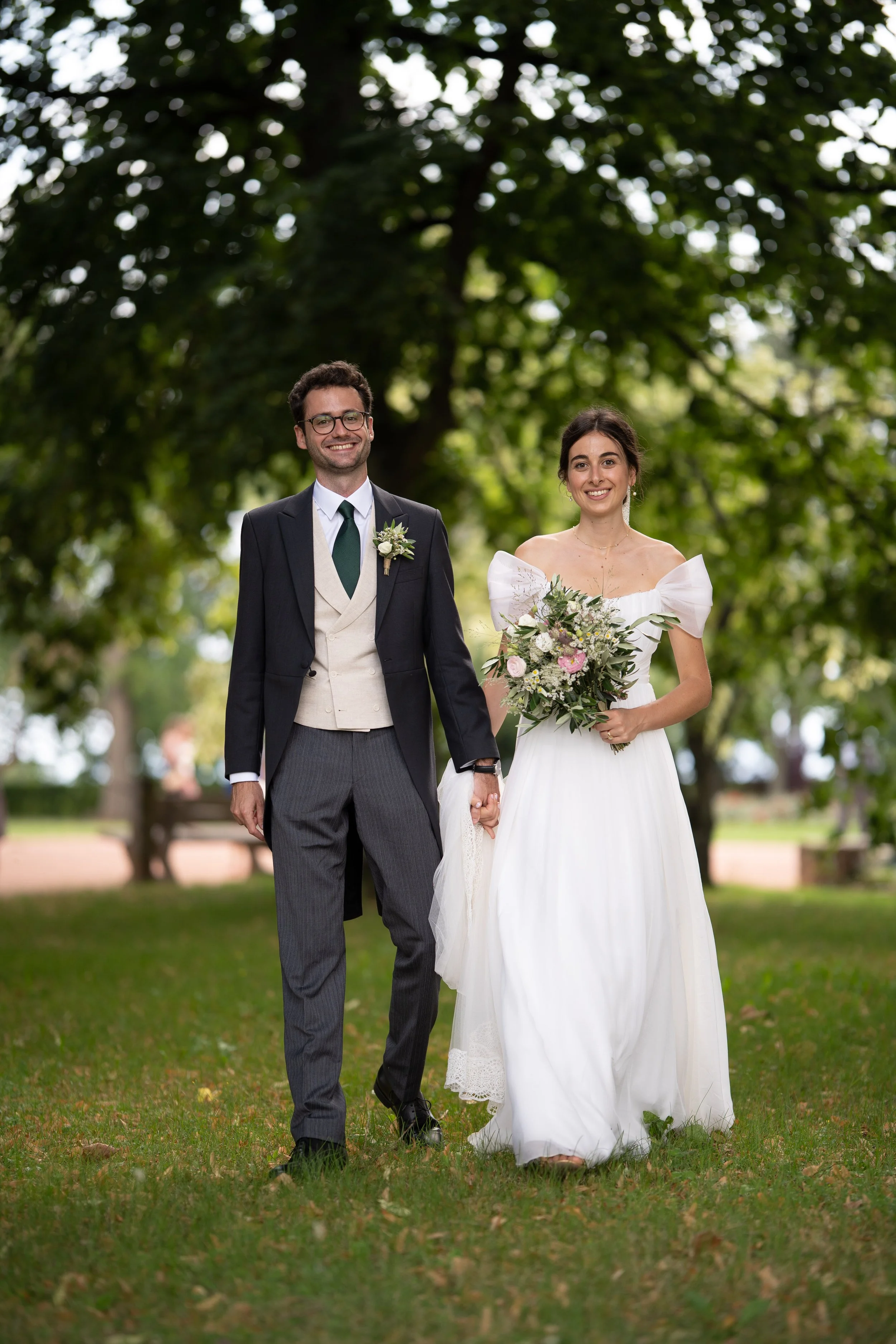 Un couple en mariage marchant dans un parc, femme portant une robe blanche avec bouquet et homme portant un costume formel.
