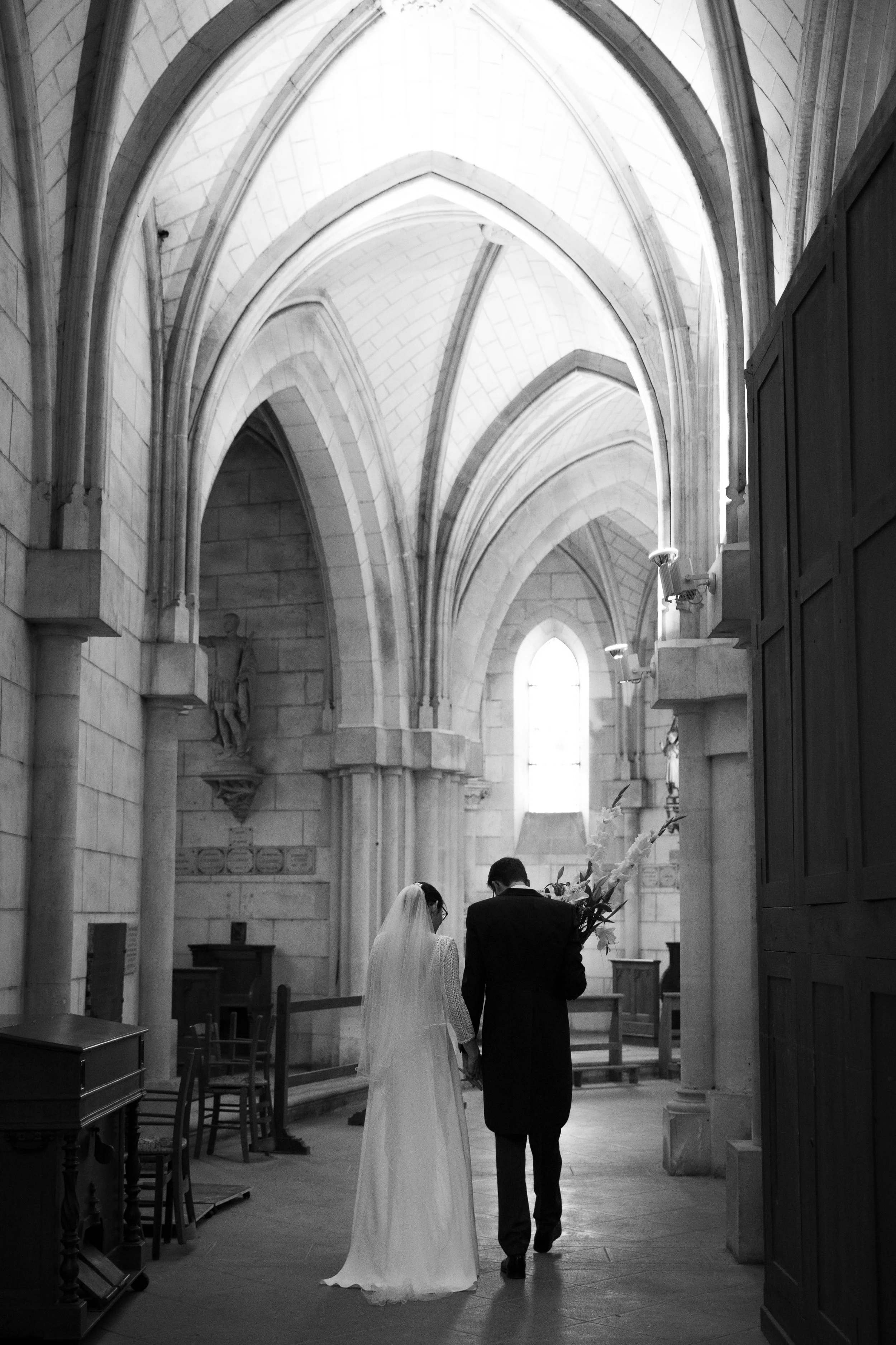 Un couple se tenant la main dans une église gothique, probablement lors d'un mariage, avec des arches en pierre et une grande fenêtre derrière eux.