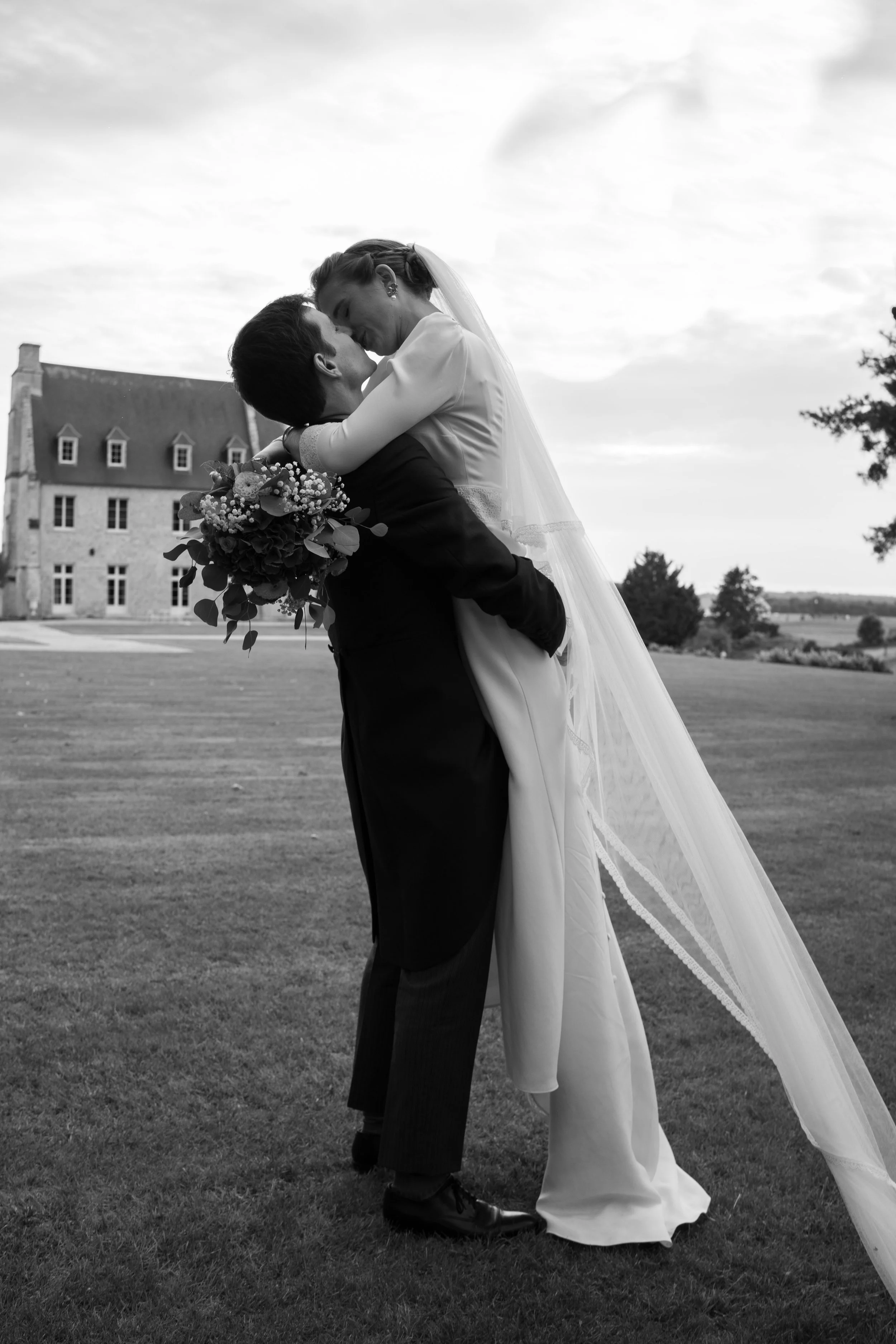 Un couple de mariés en noir et blanc, la femme en robe de mariage et l'homme en costume, se tenant dans une pose romantique dans un jardin avec une grande maison en arrière-plan.