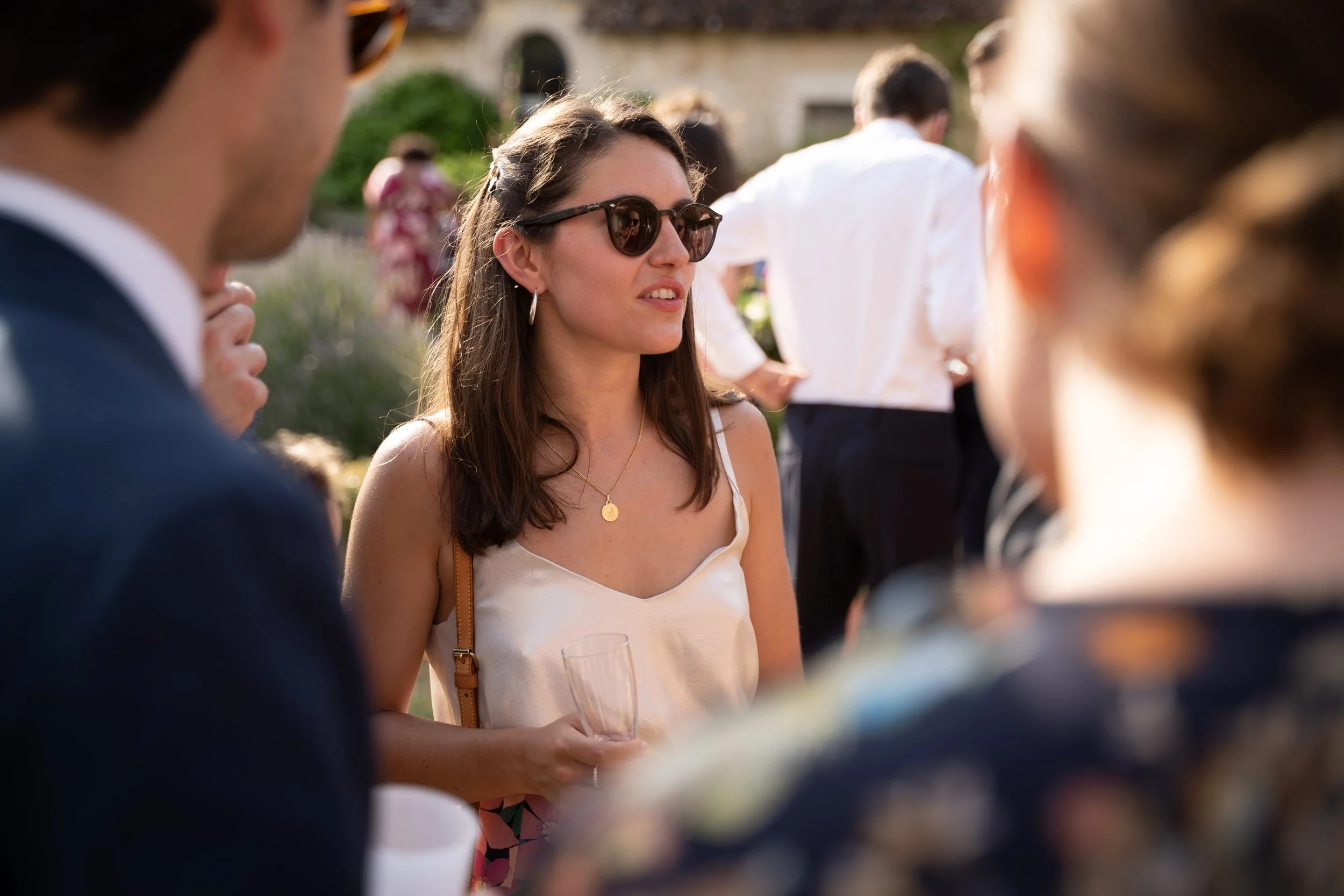 Une femme portant des lunettes de soleil et un haut blanc tient un verre à une fête en extérieur, entourée d'autres personnes dans un cadre en plein air, avec une maison en pierre en arrière-plan.