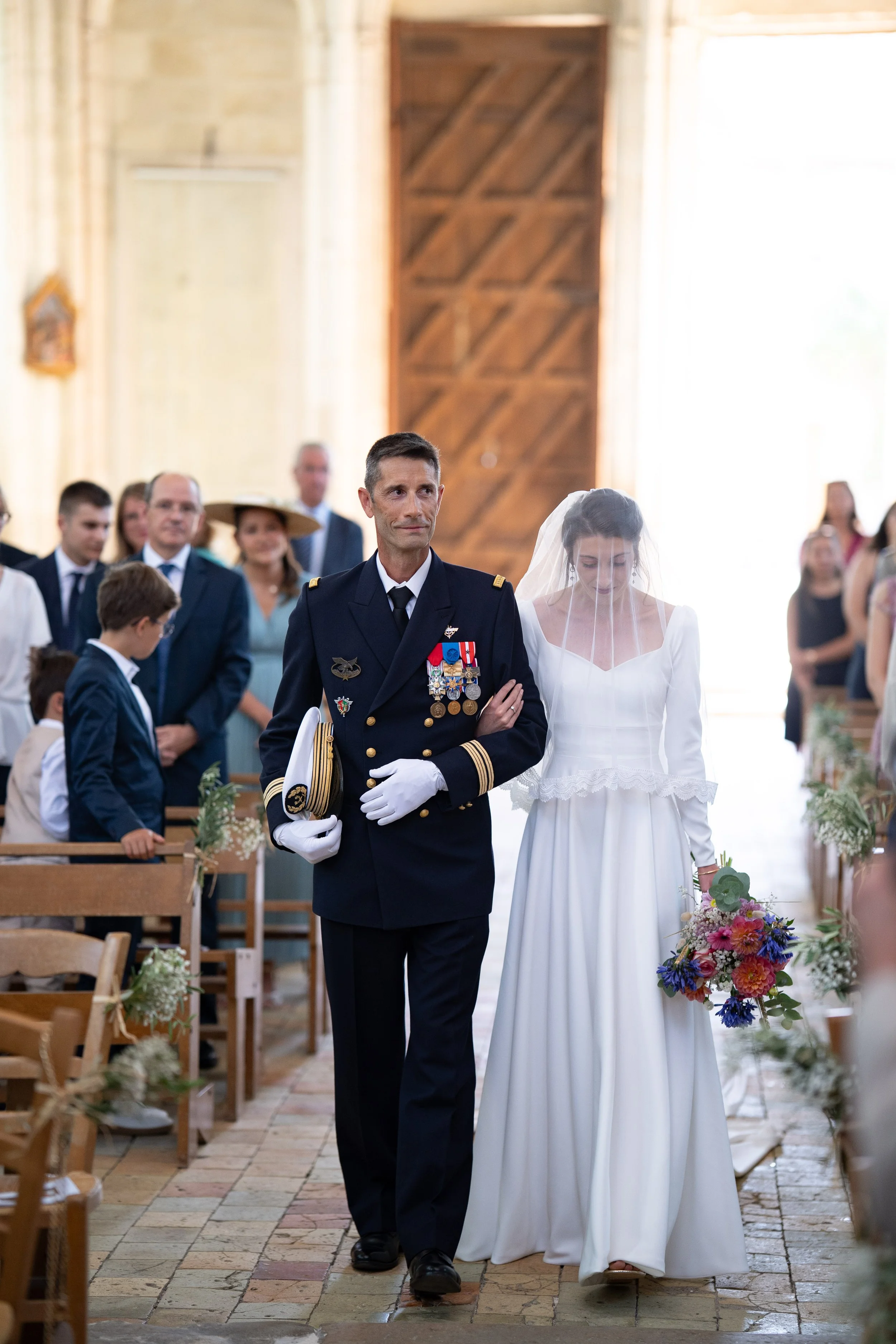 Un homme en uniforme militaire portant des médailles marche dans une cérémonie religieuse, accompagné d'une femme en robe blanche avec un voile, dans une église décorée avec des fleurs.