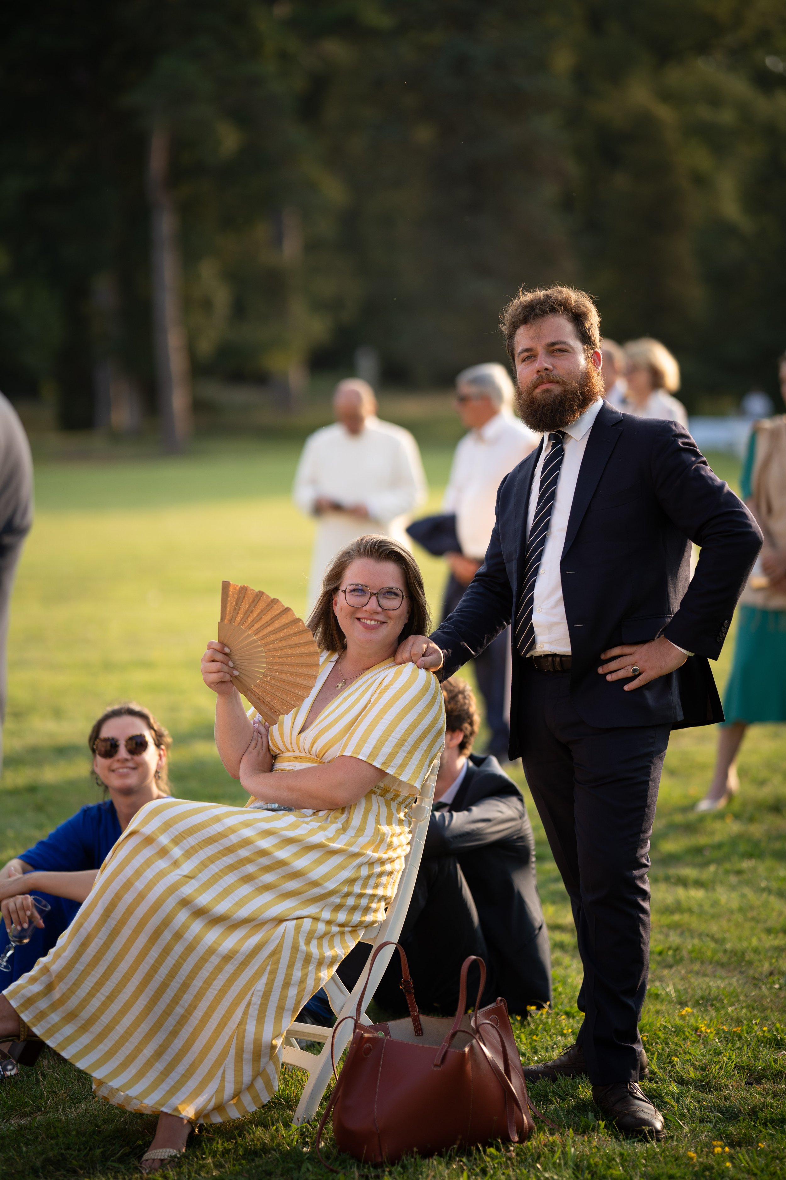 Groupe de personnes lors d'un événement en plein air, avec une femme en robe jaune à rayures assise en avant, tenant un éventail, et un homme en costume noir à côté d'elle.