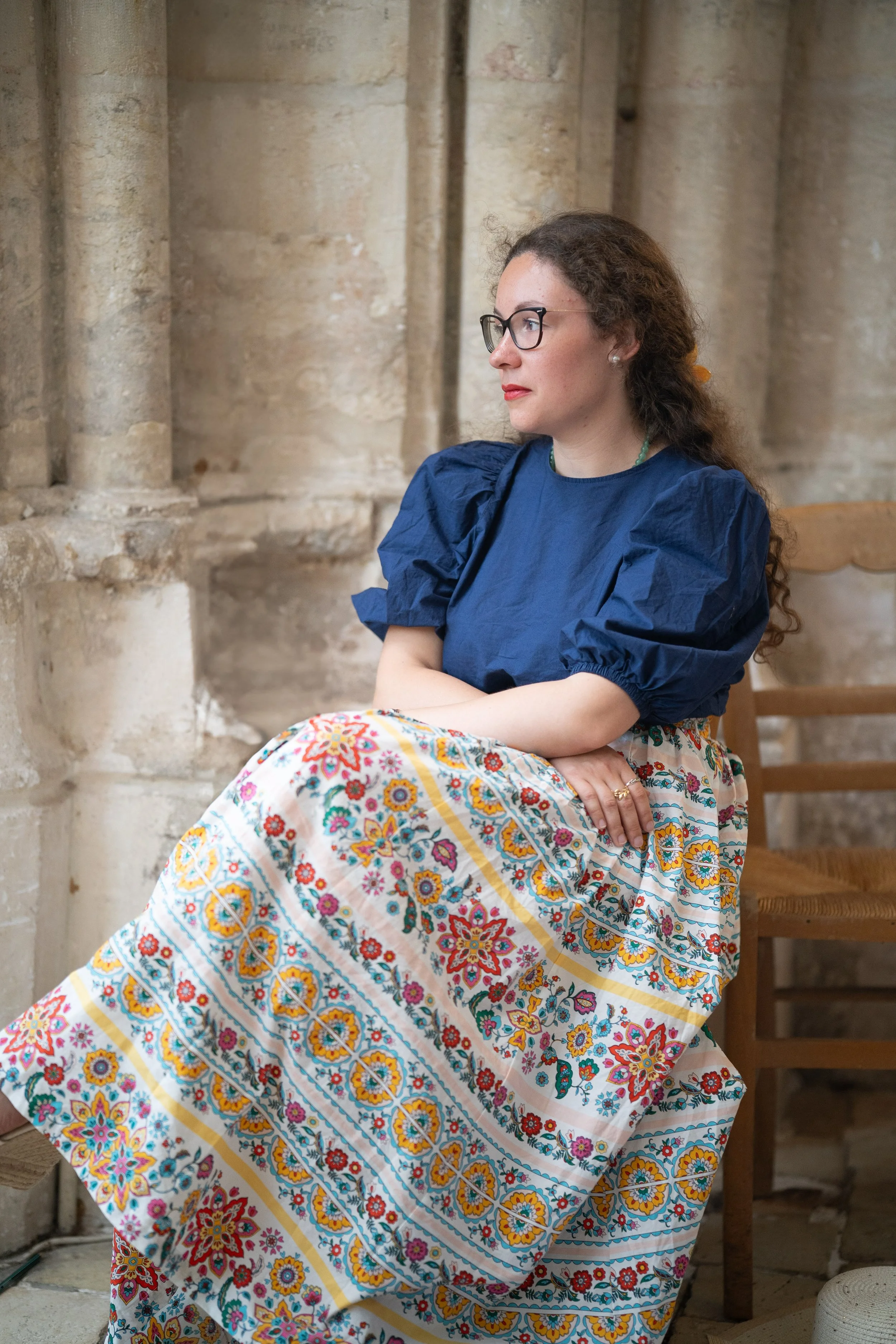 Femme assise à côté d'une colonne en pierre dans un lieu historique, portant une robe colorée avec un motif floral, lunettes, cheveux bouclés, regardant vers la droite.