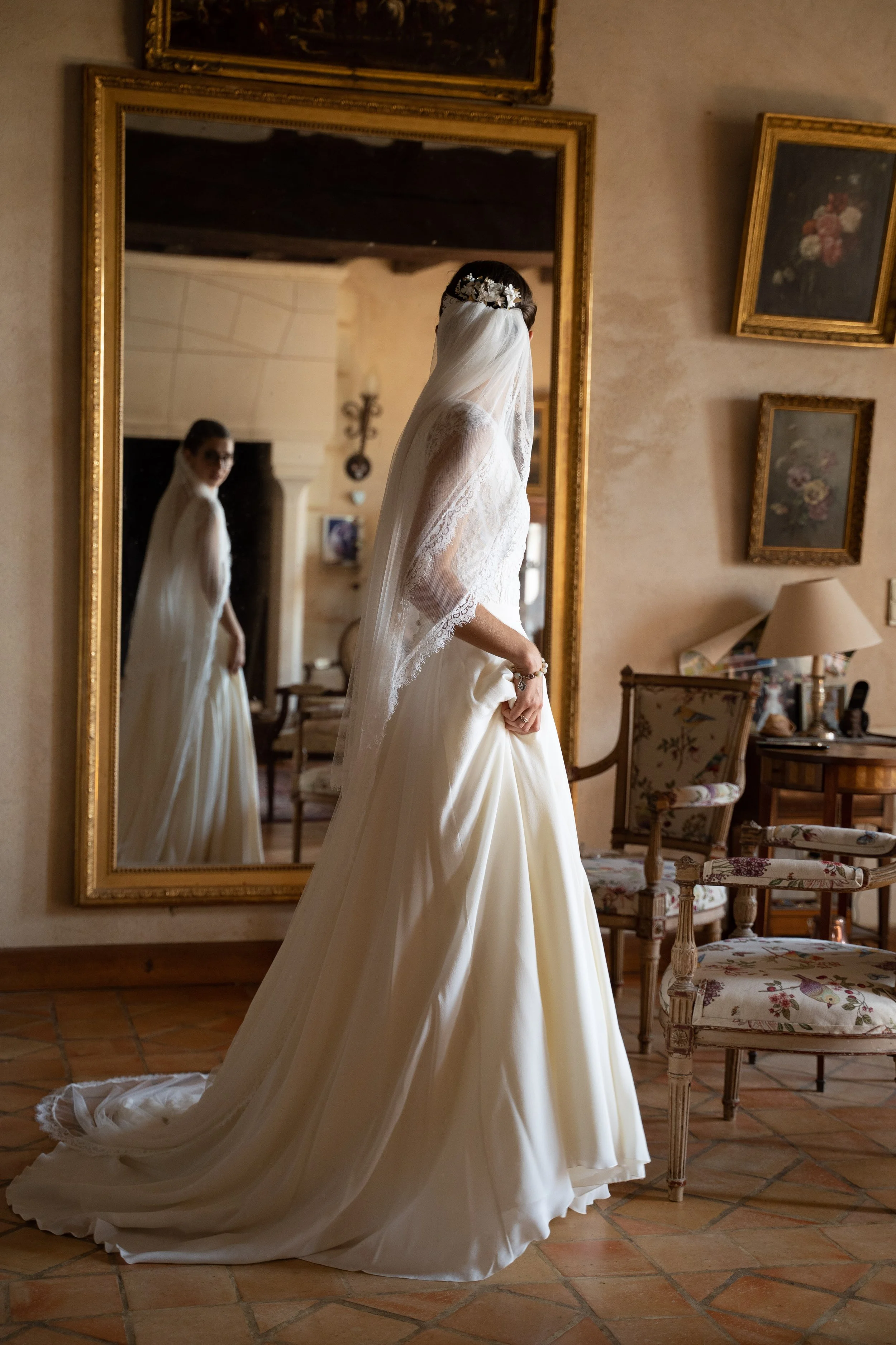 Une mariée portant une robe blanche avec un voile, vue de derrière dans un miroir ancien dans une pièce décorée avec des cadres de peintures et des meubles en bois, sol en carreaux de terre cuite.