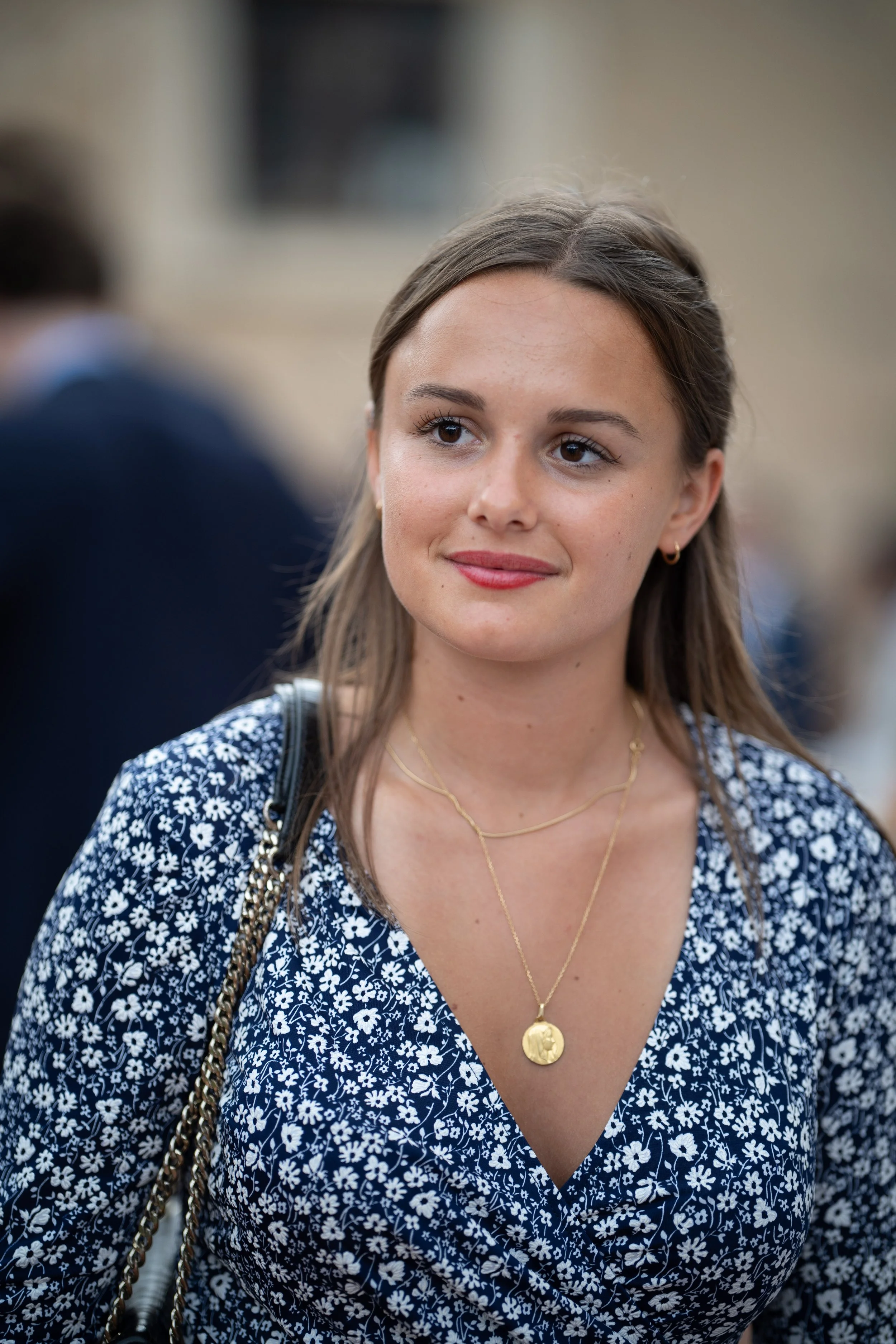 Une jeune femme avec des cheveux bruns, portant une robe à motifs floraux blanc et bleu, des bijoux en or et une chaîne avec une médaille, sourit légèrement lors d'un événement en plein air.