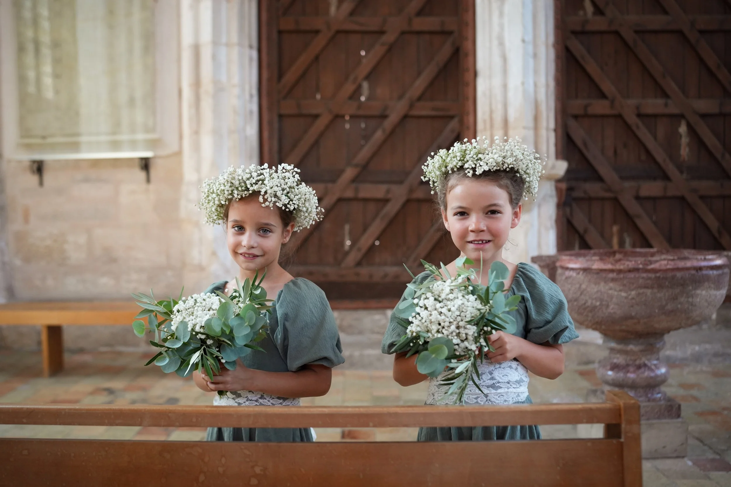 Deux jeunes fillettes portant des couronnes de fleurs blanches et tenant des bouquets de fleurs vertes se tiennent dans une église en bois, lors d'une cérémonie ou d'un événement spécial.