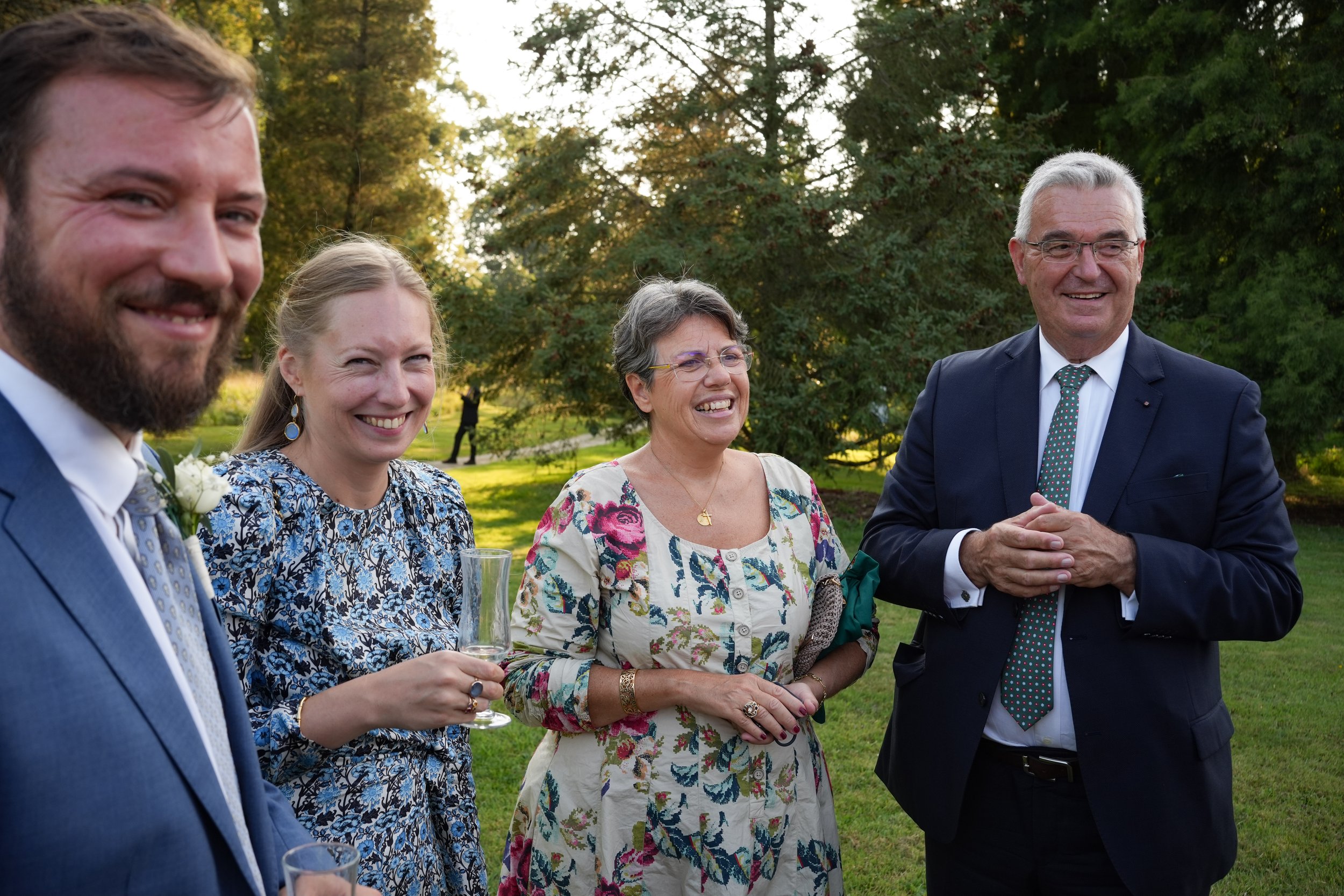 Groupe de cinq personnes à une fête en plein air, souriantes, avec des arbres en arrière-plan, certains tenant des verres à champagne.