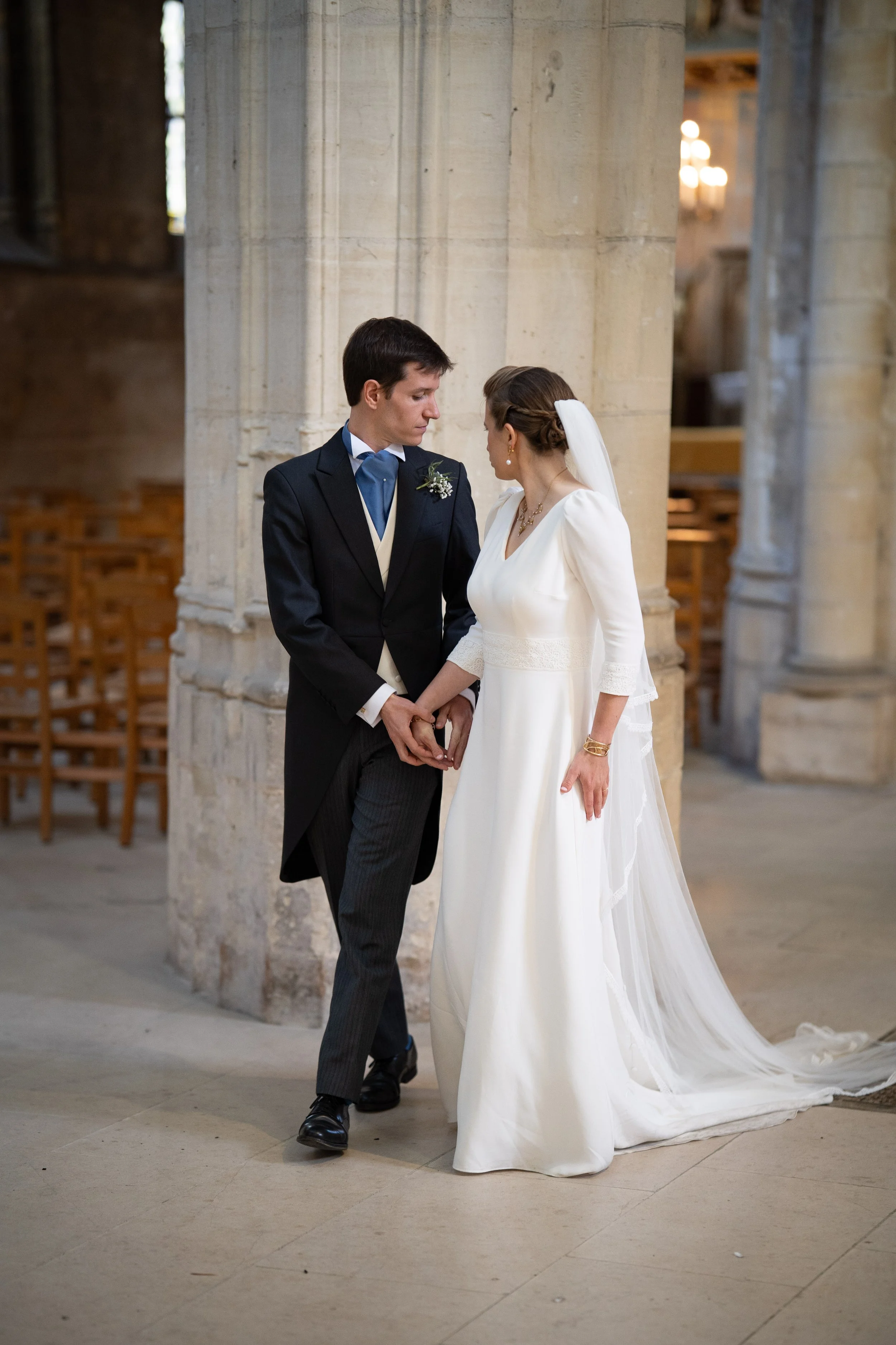 Un couple en habits de mariage se tenant la main dans une église avec des piliers en pierre et des chaises en arrière-plan.