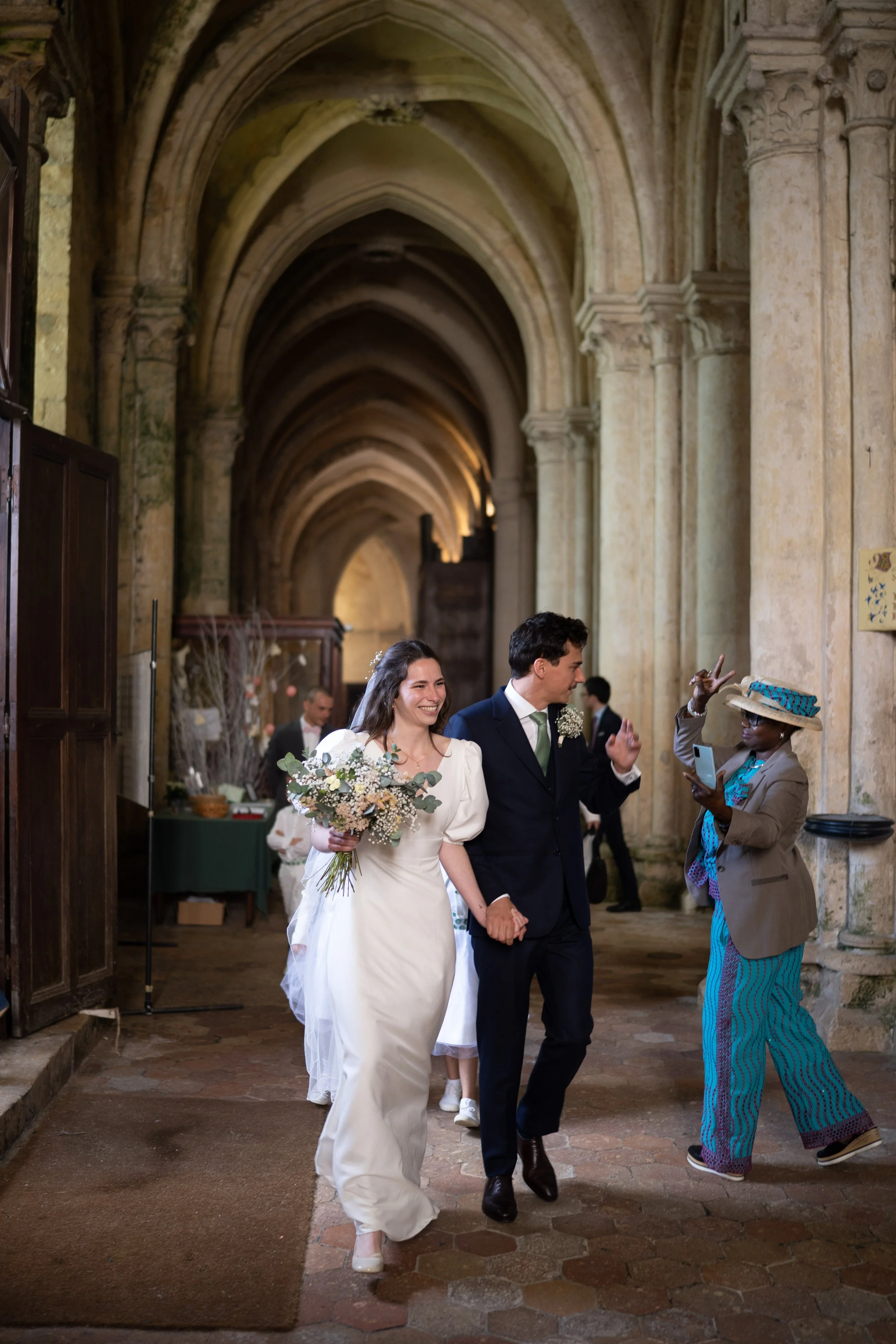 Un couple marié marchant dans une église ancienne, salué par une femme portant un chapeau coloré.