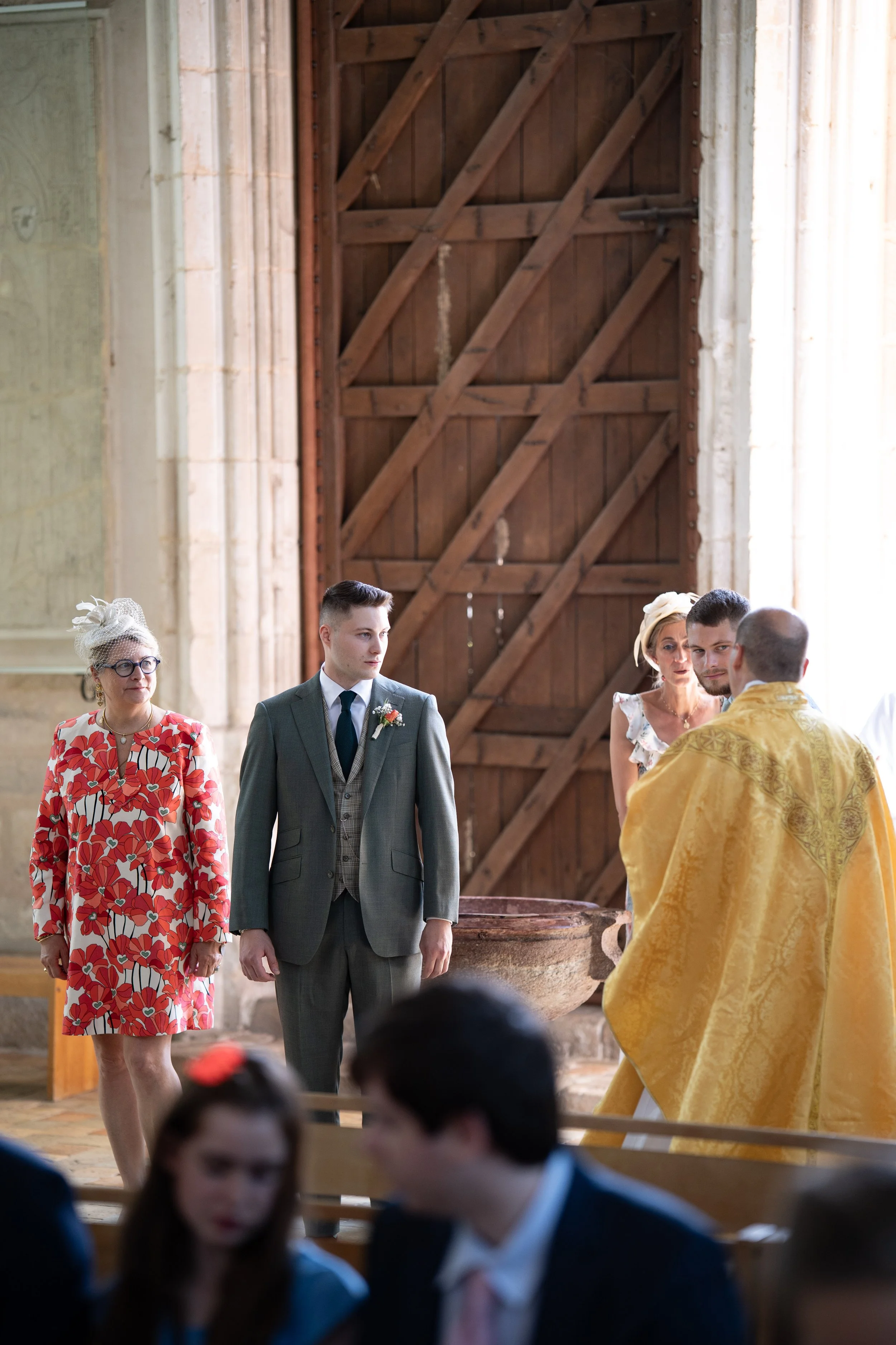 Groupe de personnes lors d'une cérémonie dans une église, avec un homme en costume, une femme en robe à motifs rouges et blancs, et d'autres personnes en arrière-plan, dans un cadre religieux en pierre et bois.