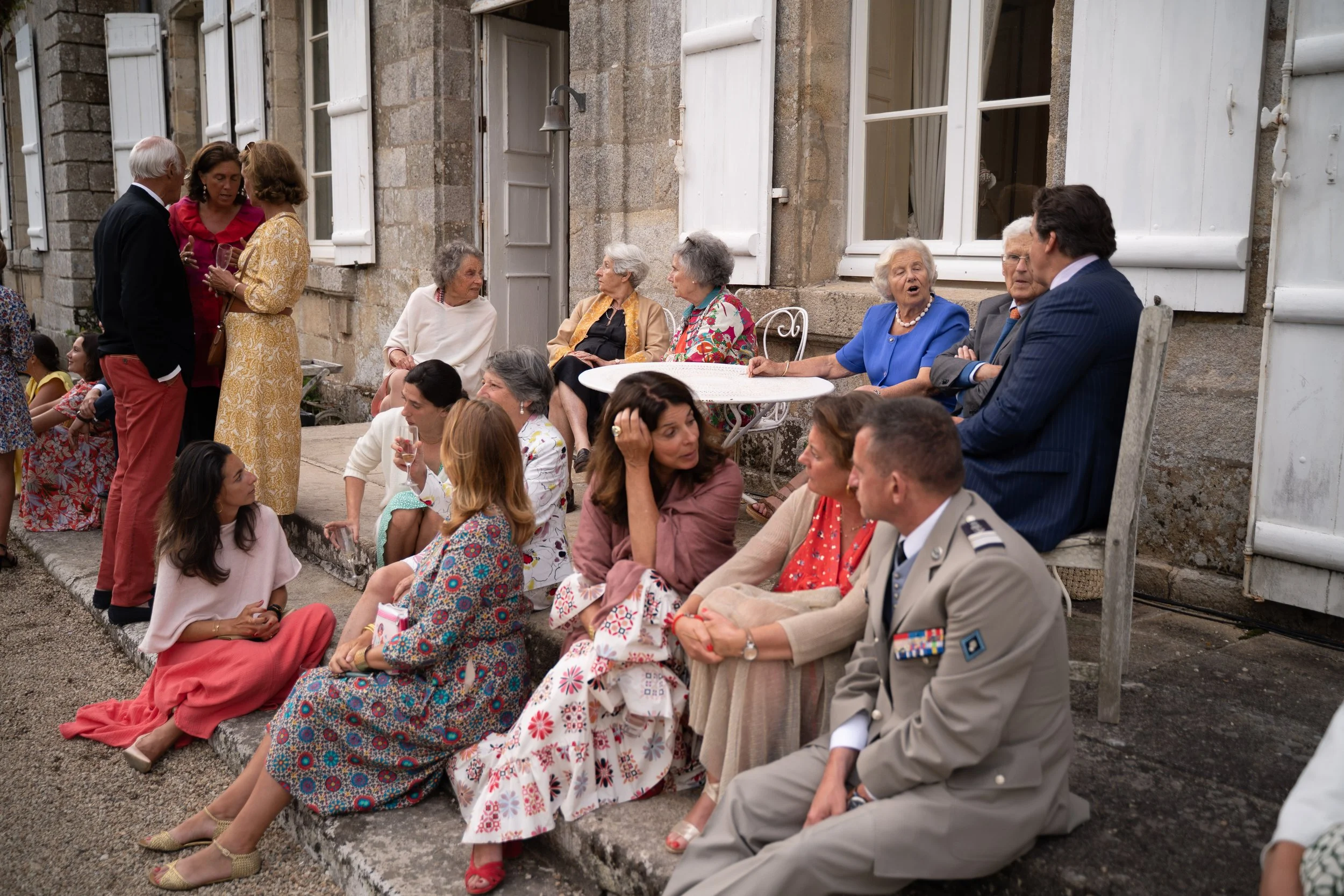 Groupe de personnes âgées et jeunes assises ou discutant devant une vieille maison en pierres, avec fenêtres à volets blancs, lors d'un rassemblement en plein air.