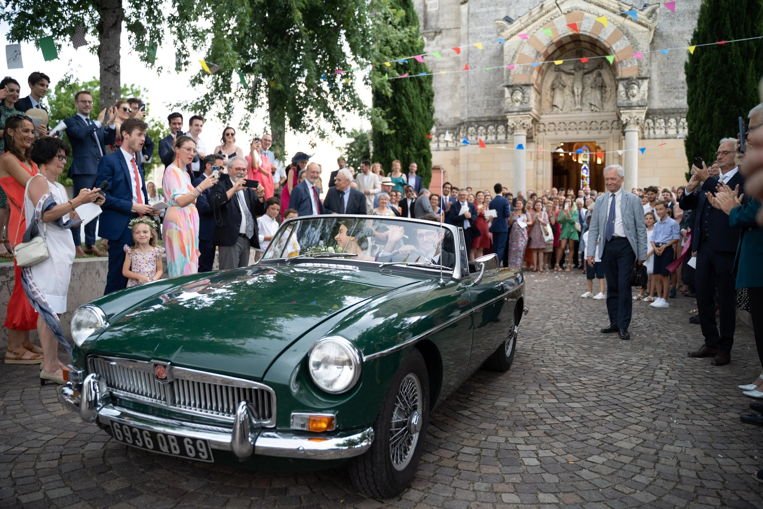 Une foule de personnes habillées élégamment assiste à une célébration en extérieur devant une église en pierre, avec des drapeaux colorés en décoration, et une voiture classique verte au centre de la scène.