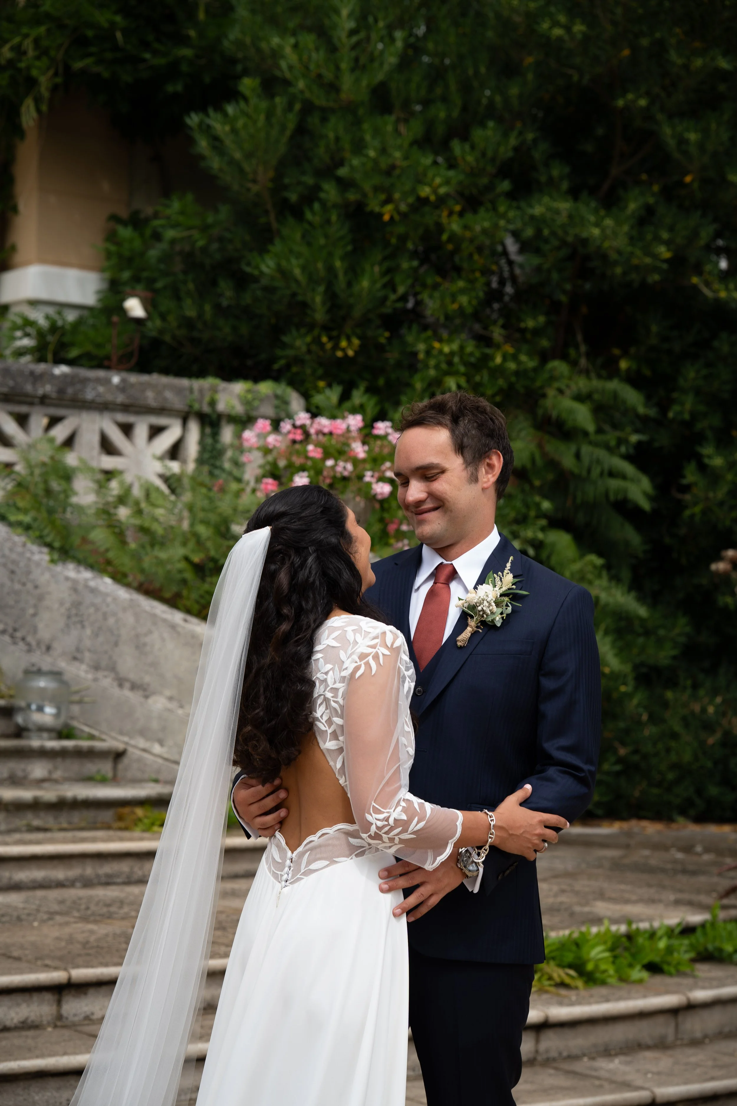 Un couple de mariés en tenue de mariage, se regardant tendrement lors d'une cérémonie en plein air, avec des fleurs roses et un jardin verdoyant en arrière-plan.