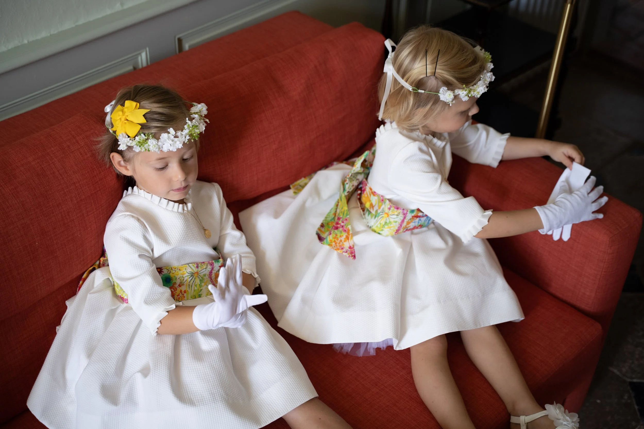 Deux jeunes filles en robes blanches, chapeaux de fleurs, assises sur un canapé rouge, avec des gants blancs, semblent à une cérémonie ou un événement formel.