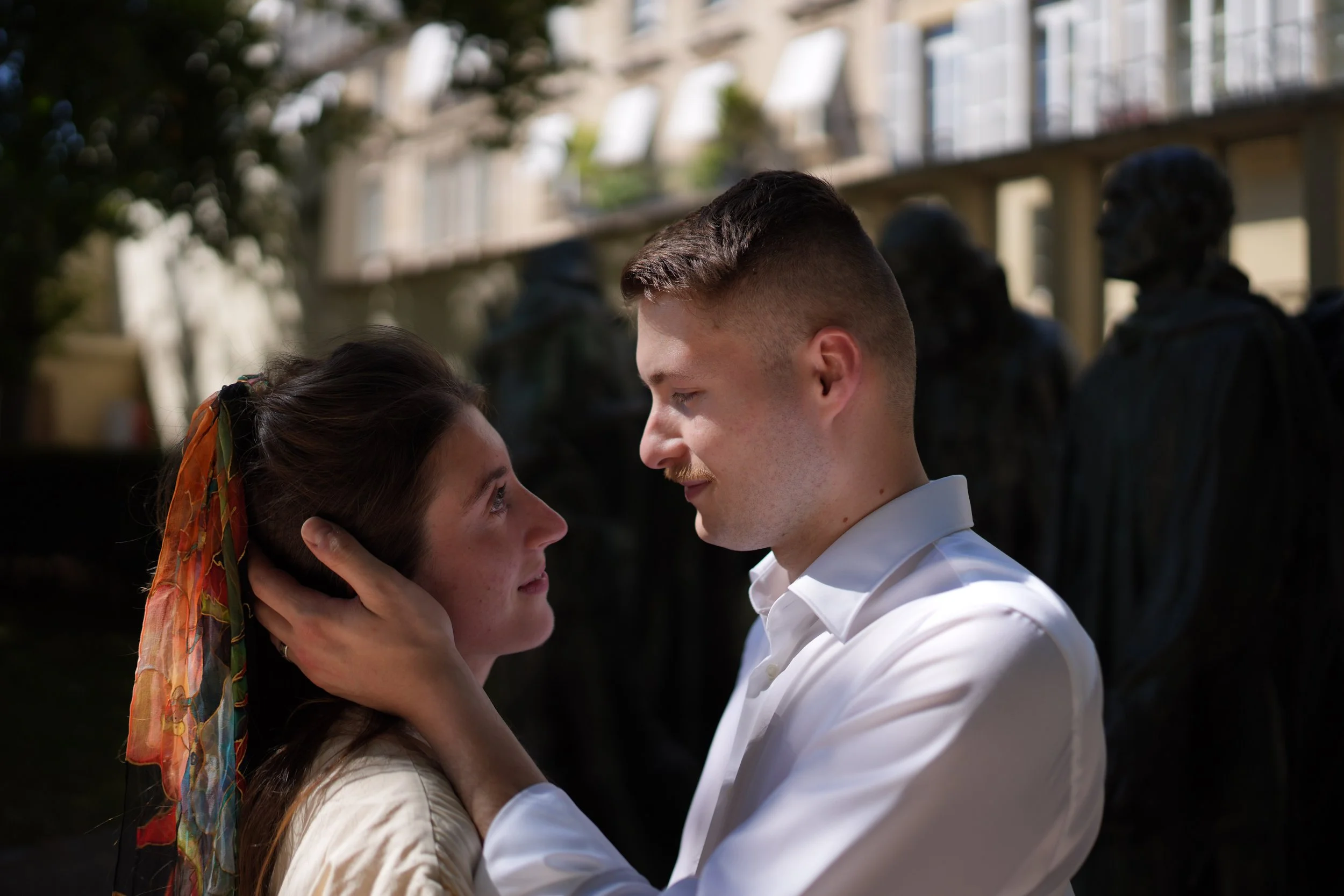 Un couple regarde tendrement l'un l'autre, un homme et une femme se tiennent face à face dehors avec un bâtiment en arrière-plan, ils sont émus et proches, créant une scène intime et romantique.