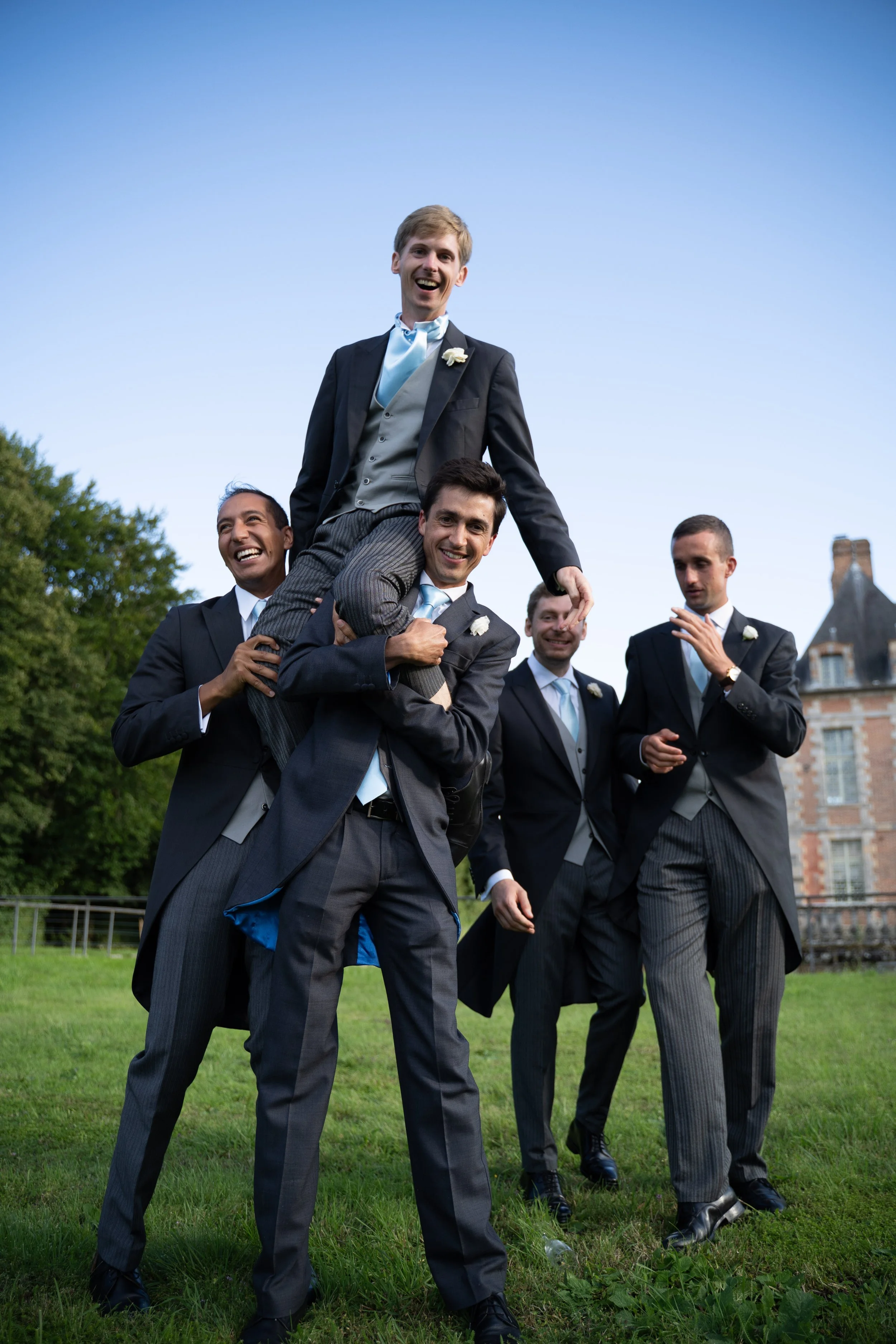 Groupe d'hommes d'affaires en costume, certains souriants, participant à une activité de célébration en extérieur, avec un bâtiment historique en arrière-plan et un ciel dégagé.