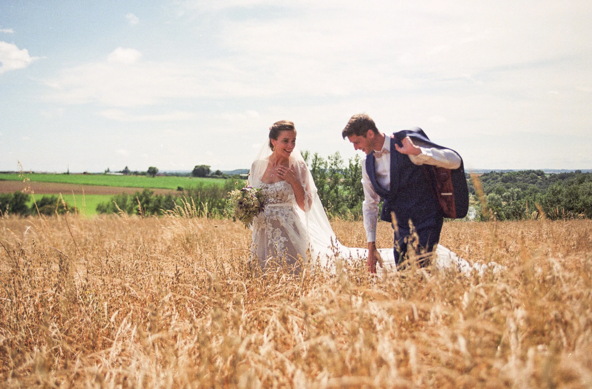 Un couple en mariage se promenant dans un champ de blé doré sous un ciel bleu avec quelques nuages, dans un paysage rural verdoyant.