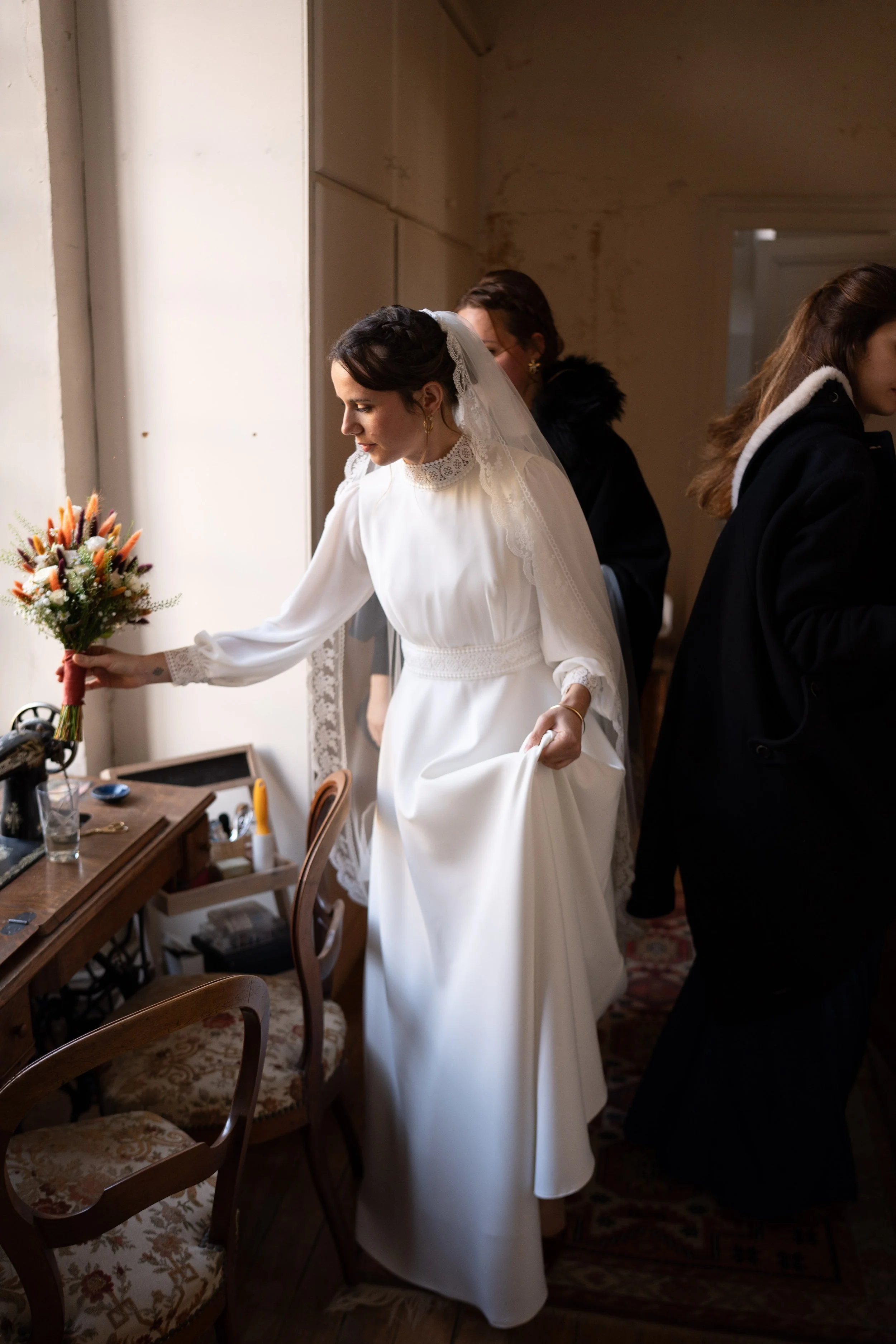 Une mariée en robe blanche avec un voile, tenant un bouquet de fleurs, se préparant avec l'aide de deux autres femmes dans une pièce