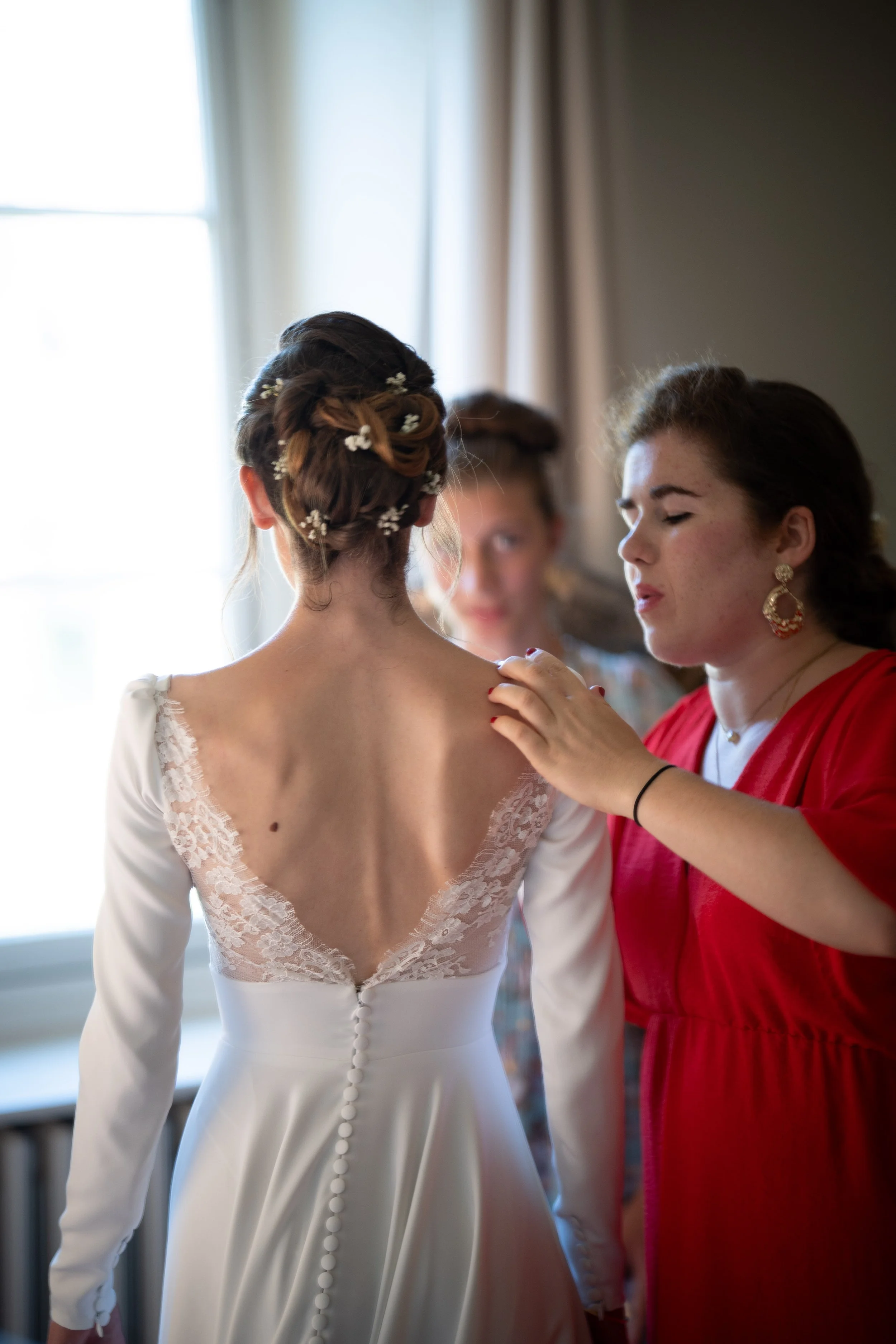 Une femme en robe de mariée avec des boutons dans le dos, une autre femme en robe rouge ajuste ou vérifie la robe de la mariée, dans une pièce lumineuse