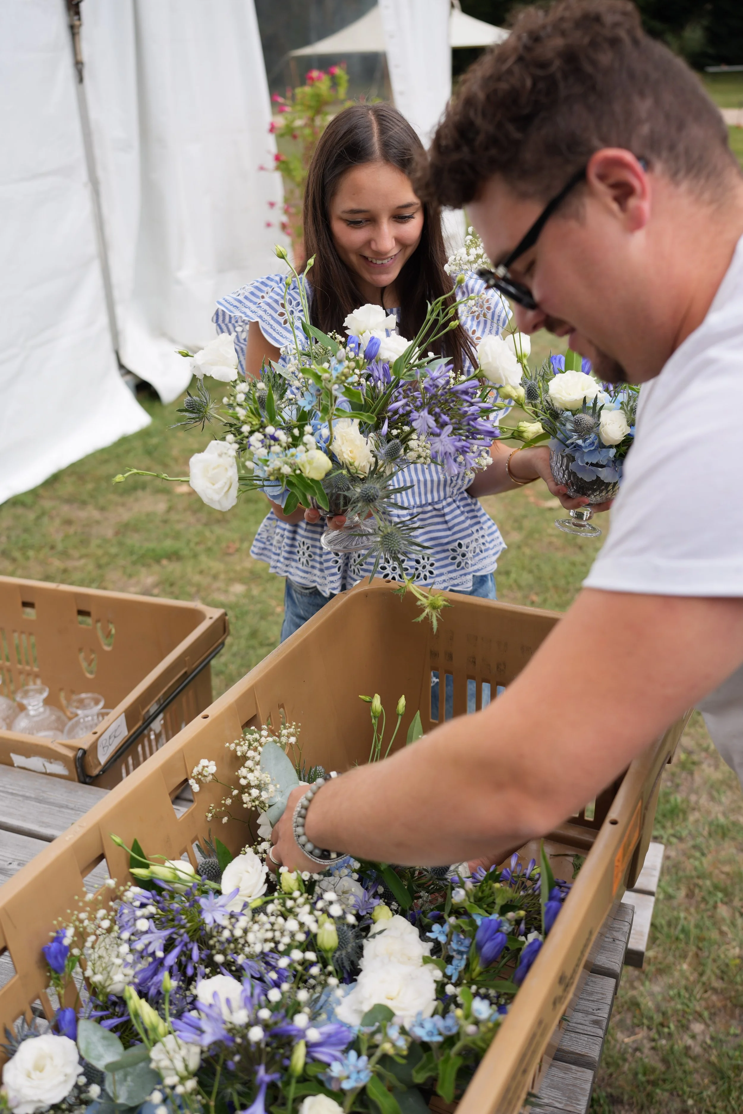 Deux personnes, une femme et un homme, trient des fleurs dans des paniers en plastique lors d'un événement en plein air. La femme sourit et porte une robe à rayures bleues et blanches, tandis que l'homme porte des lunettes et un t-shirt blanc.
