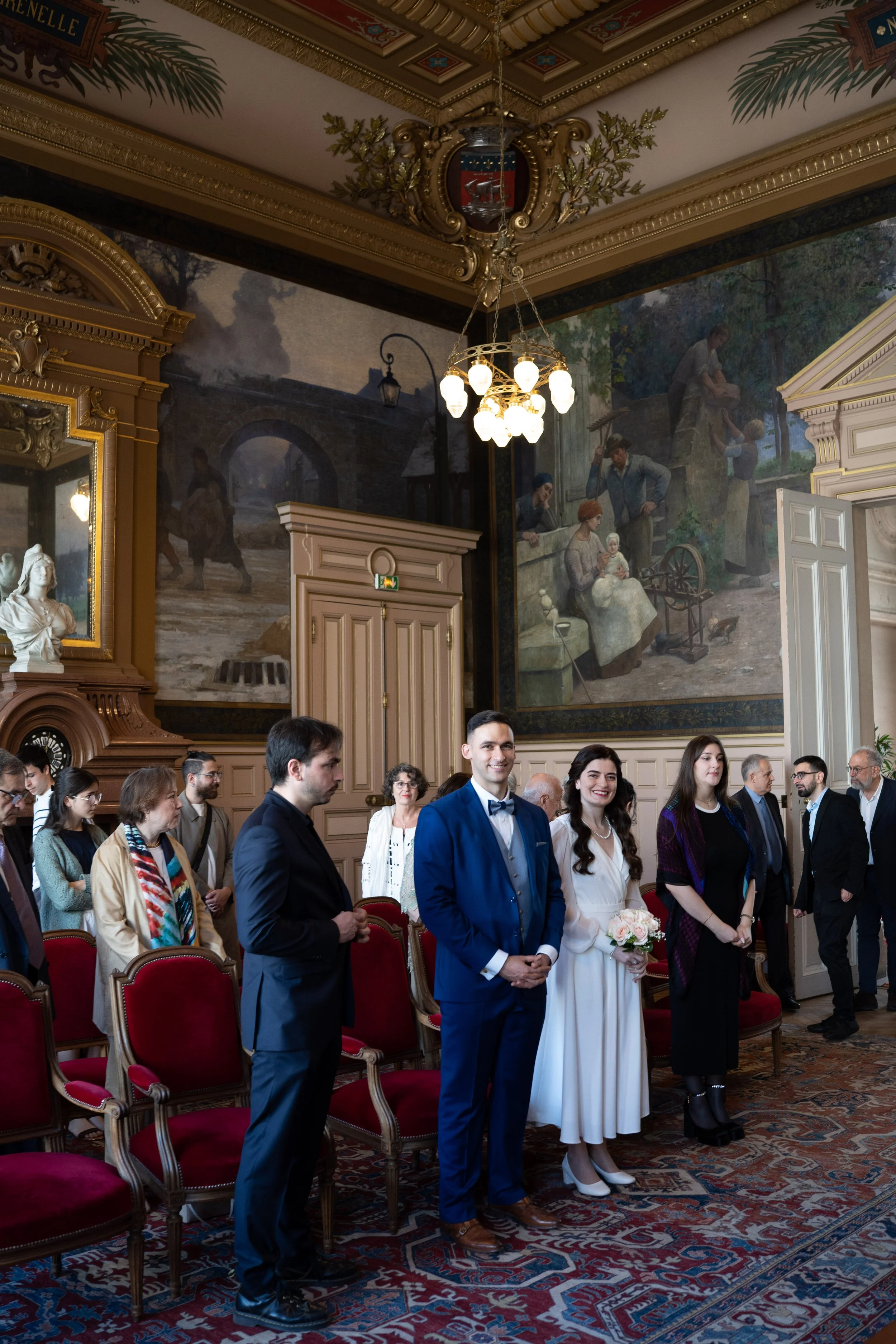 Un groupe de personnes habillées formellement lors d'une cérémonie ou d'un mariage dans une salle ornée de peintures et de meubles anciens, avec un plafond décoratif et un chandelier lumineux.