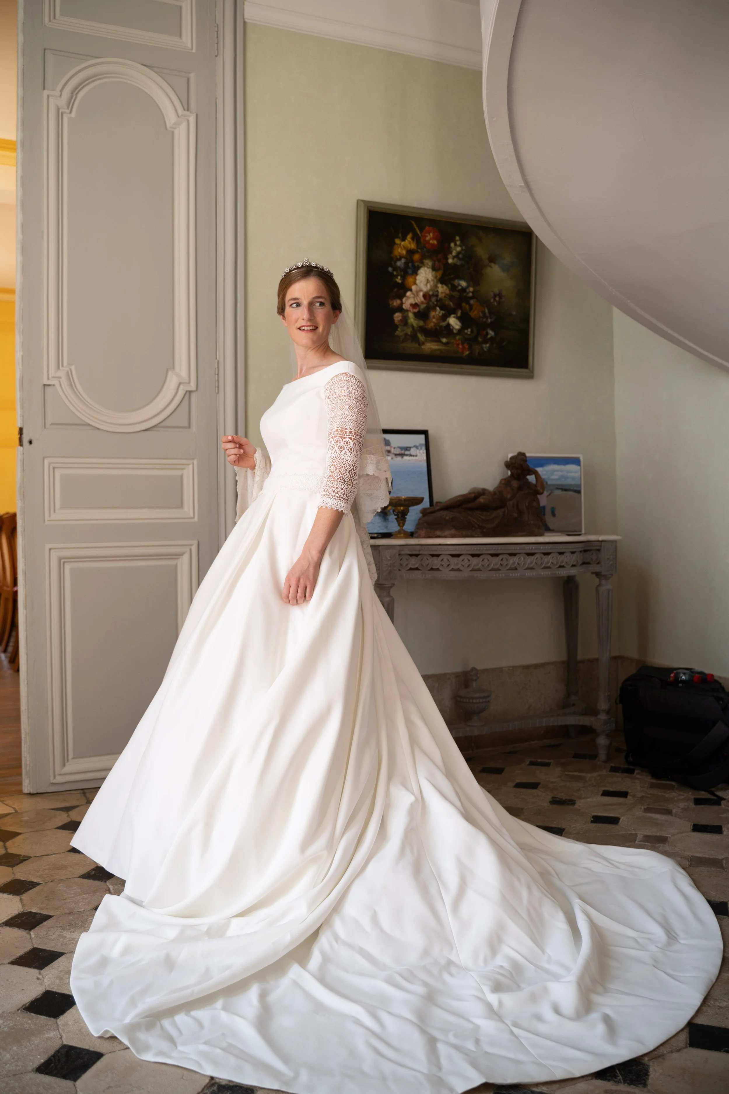 Jeune femme en robe de mariage blanche avec un diadème, dans une pièce élégante avec un tableau de fleurs, un meuble ancien et un sol en carreaux.