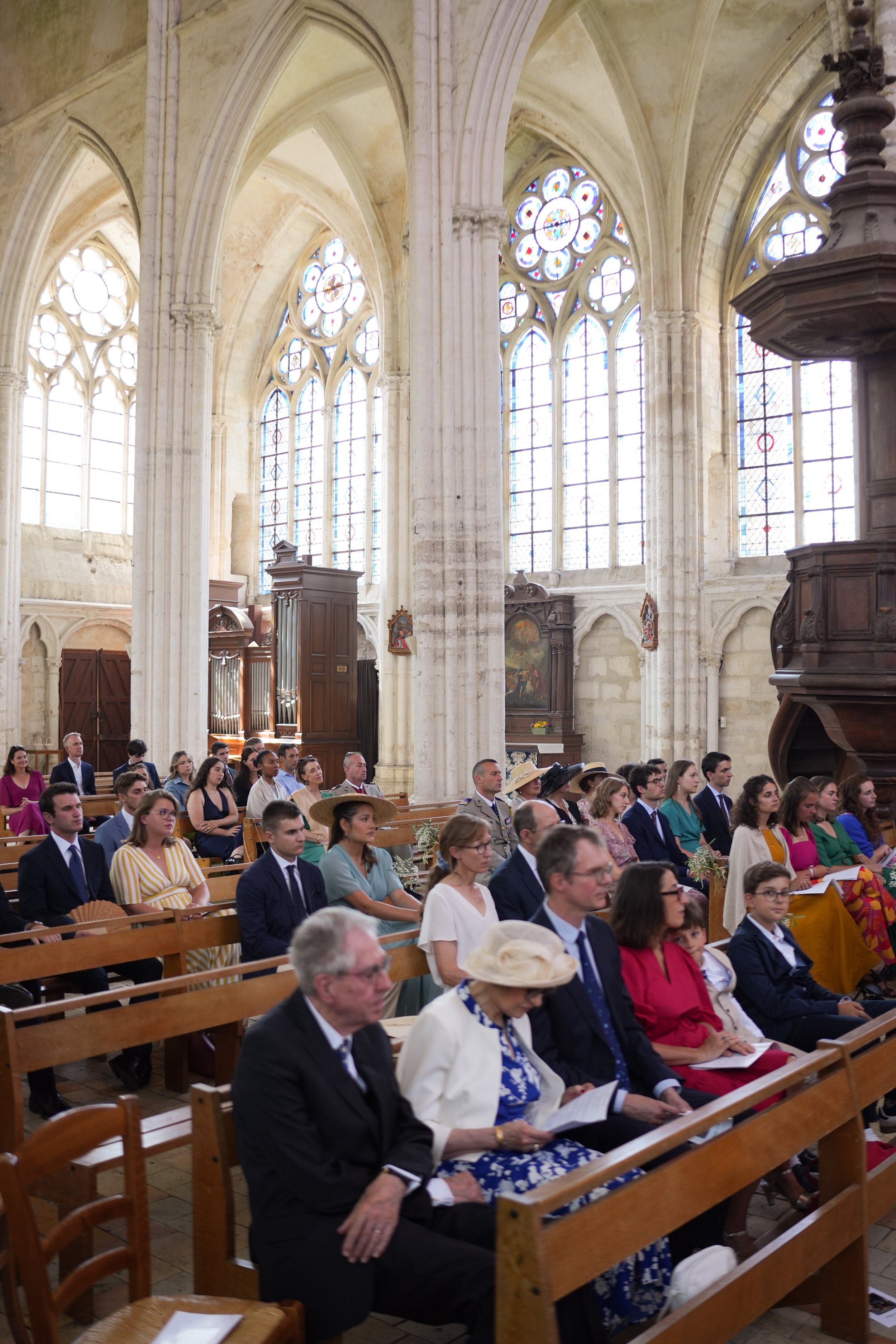 Un groupe de personnes assises dans une église gothique, vêtues de tenues formelles, assistant à une cérémonie ou une célébration religieuse, avec des vitraux colorés en arrière-plan.