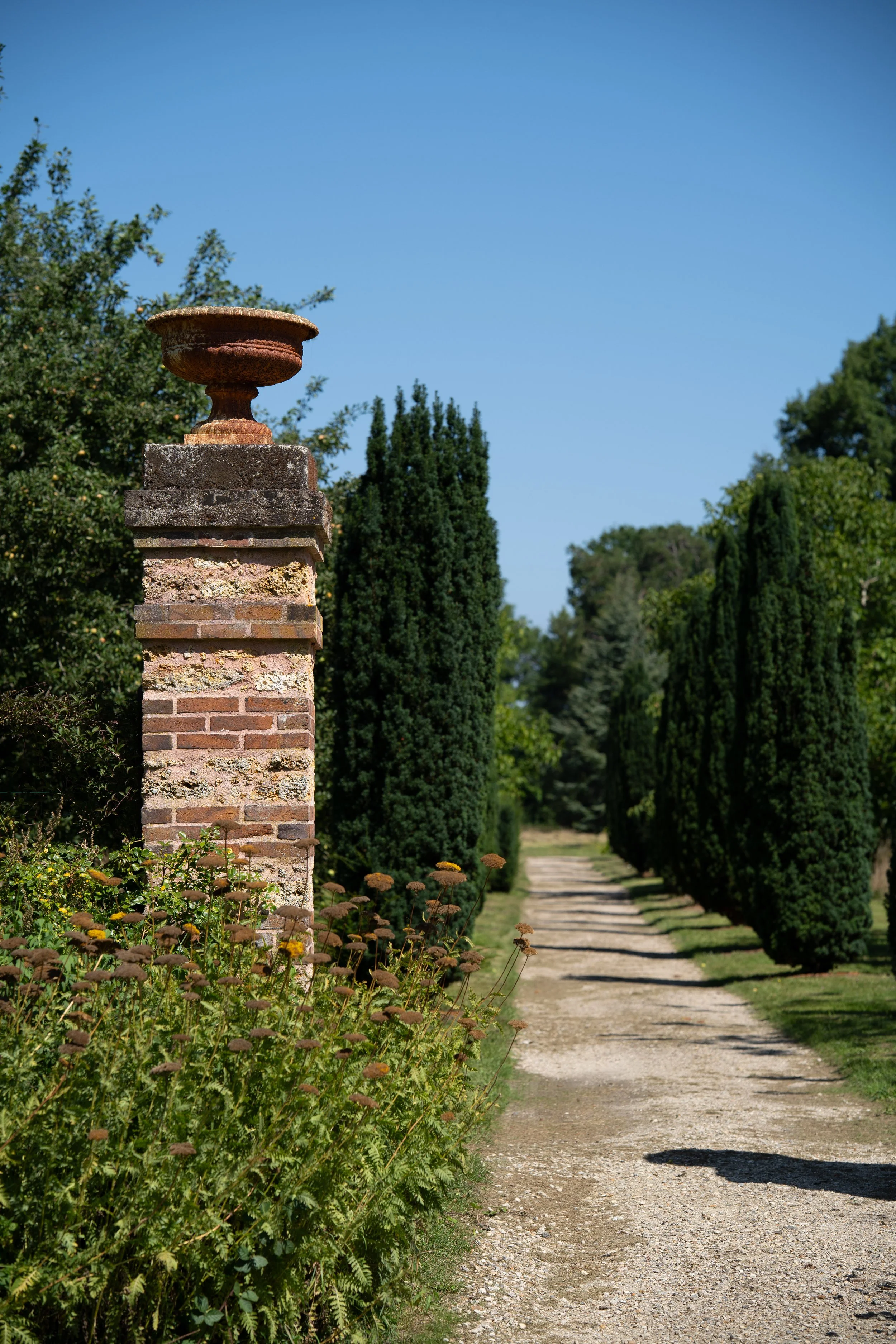 Sentier en gravier bordé d'arbres verts, avec une vieille colonne en pierre et une jarre en terre cuite sur une murette en briques, sous un ciel bleu clair.