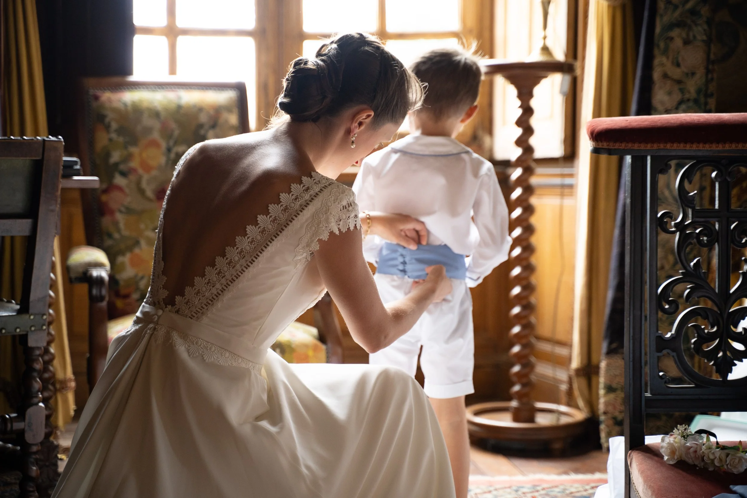 Une femme en robe de mariée inclinée, touchant le sol, dans une pièce bien éclairée par la lumière naturelle provenant d'une grande fenêtre. Deux enfants, l'un avec une chemise blanche et l'autre avec une tenue blanche et bleue, se tiennent derrière 