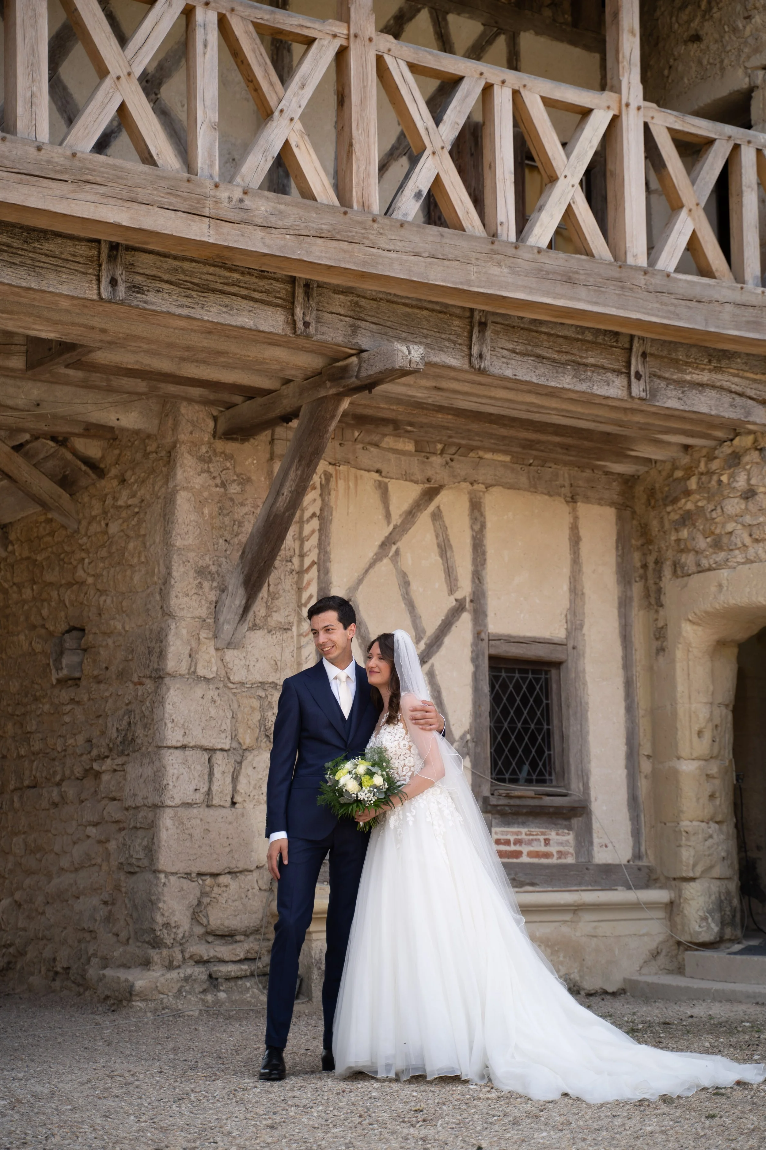 Un couple de mariés posant devant un bâtiment en pierre, la femme tenant un bouquet de fleurs blanc et vert, l'homme en costume noir, dans un décor rustique.