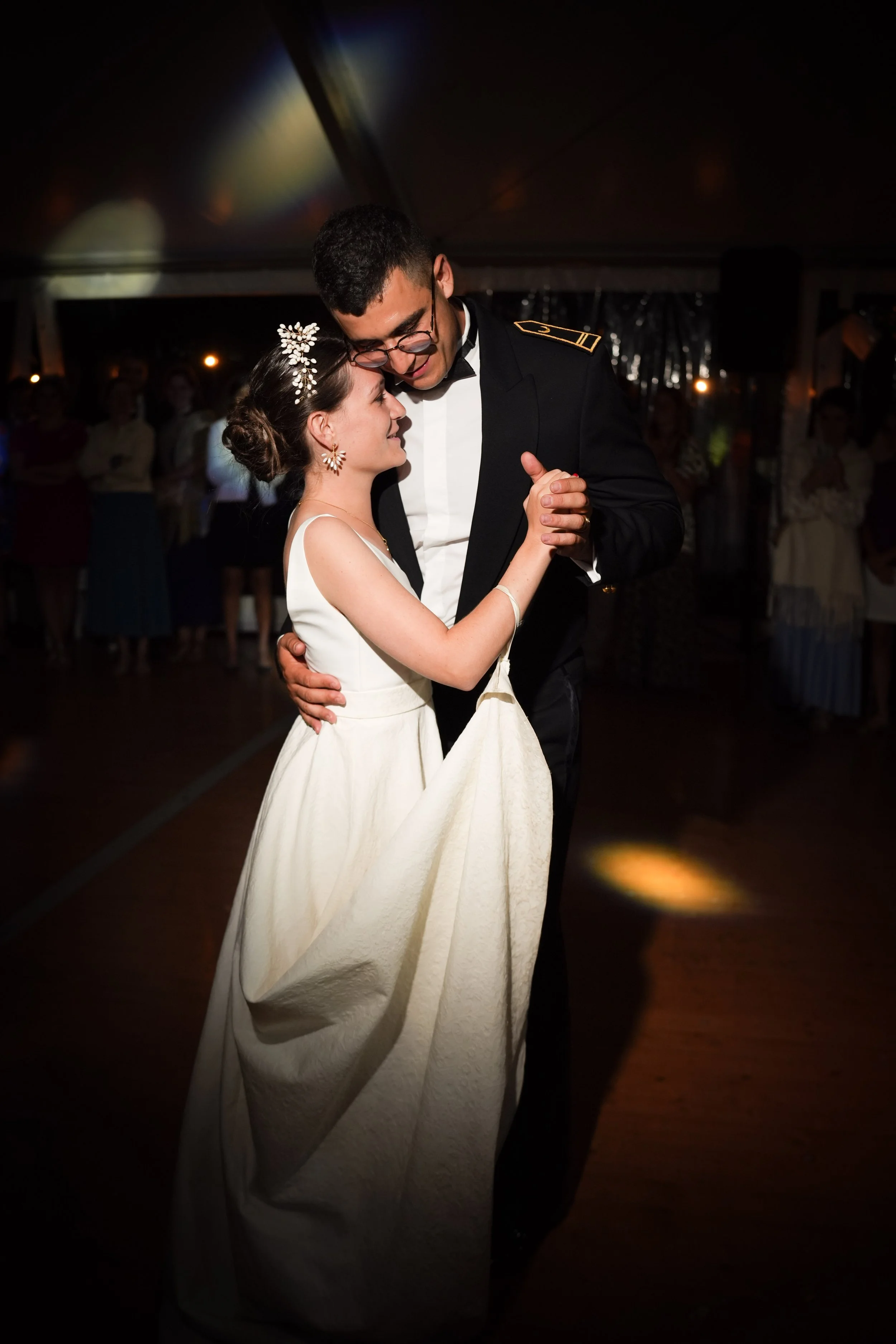 Un couple danse lors d'une cérémonie ou d'une fête, la femme porte une robe de mariée blanche et des accessoires floraux dans les cheveux, l'homme porte un uniforme noir.