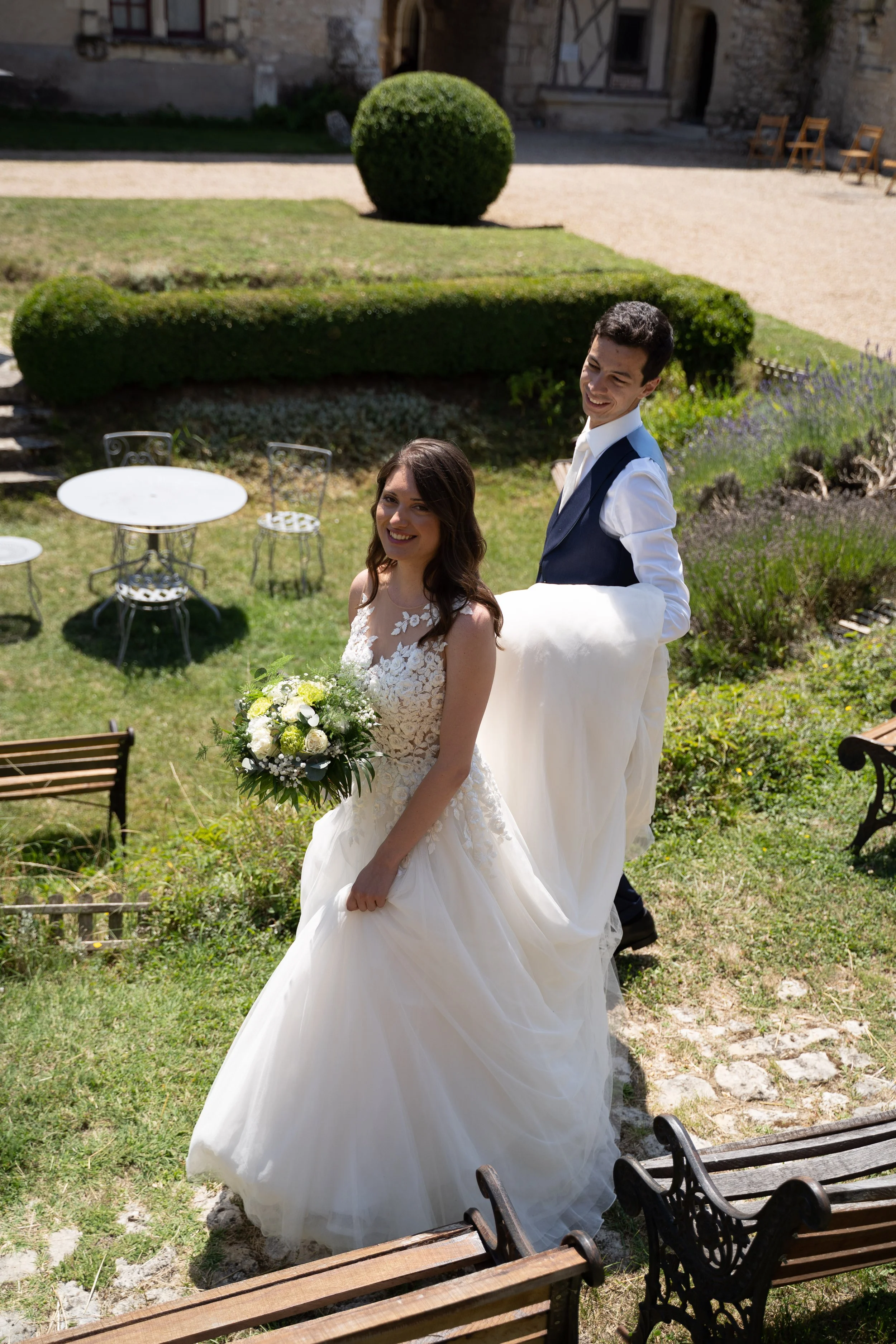 Un couple en tenue de mariage dans un jardin ensoleillé, la femme porte une robe blanche et tient un bouquet de fleurs, l'homme porte un costume avec une chemise blanche.