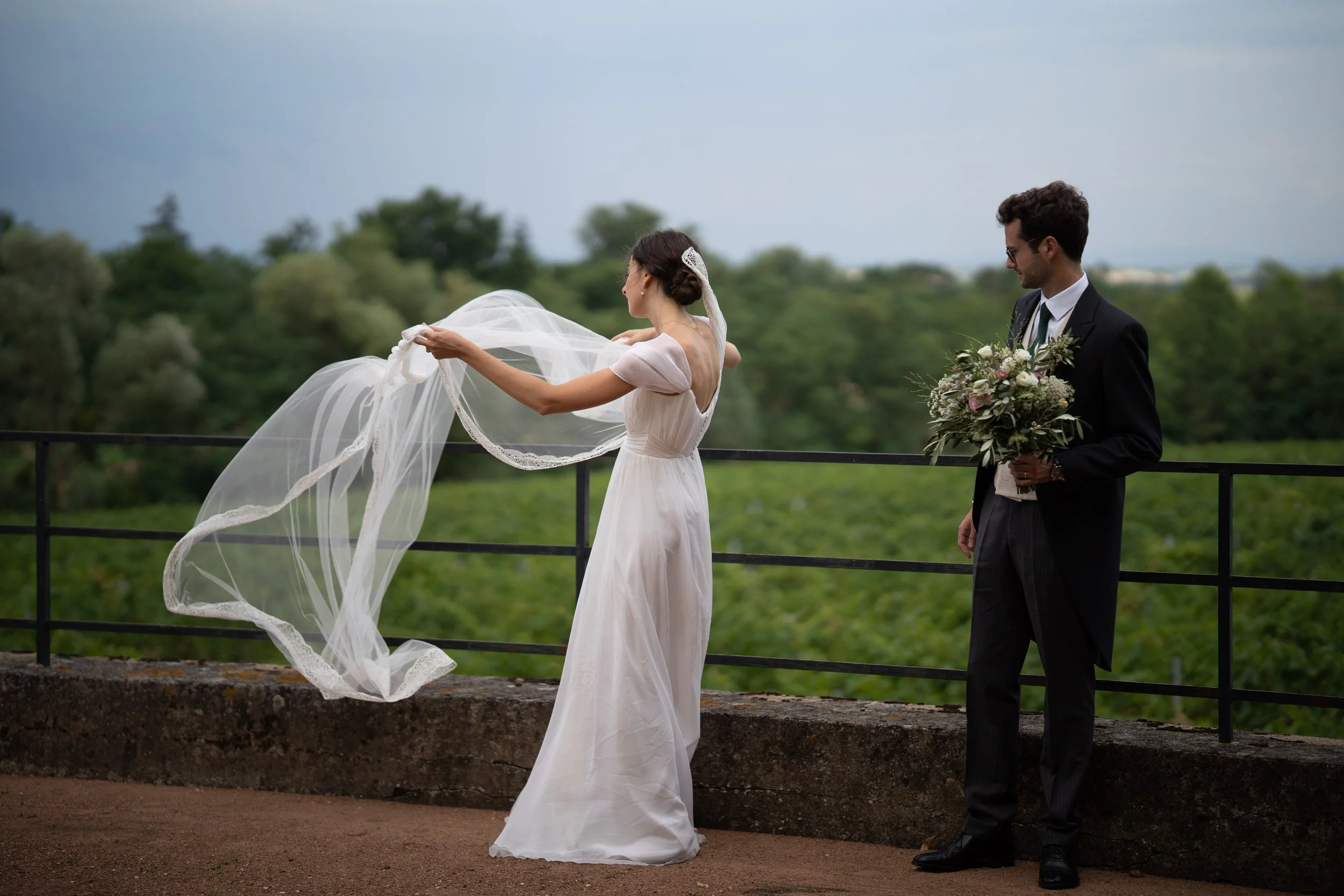 Un couple lors d'un mariage en plein air. La femme porte une robe de mariée blanche avec une longue traîne et une voilette qu'elle tient dans ses mains. L'homme porte un costume noir avec une cravate et tient un bouquet de fleurs. La scène est située sur une terrasse avec un arrière-plan de verdure et de ciel nuageux.
