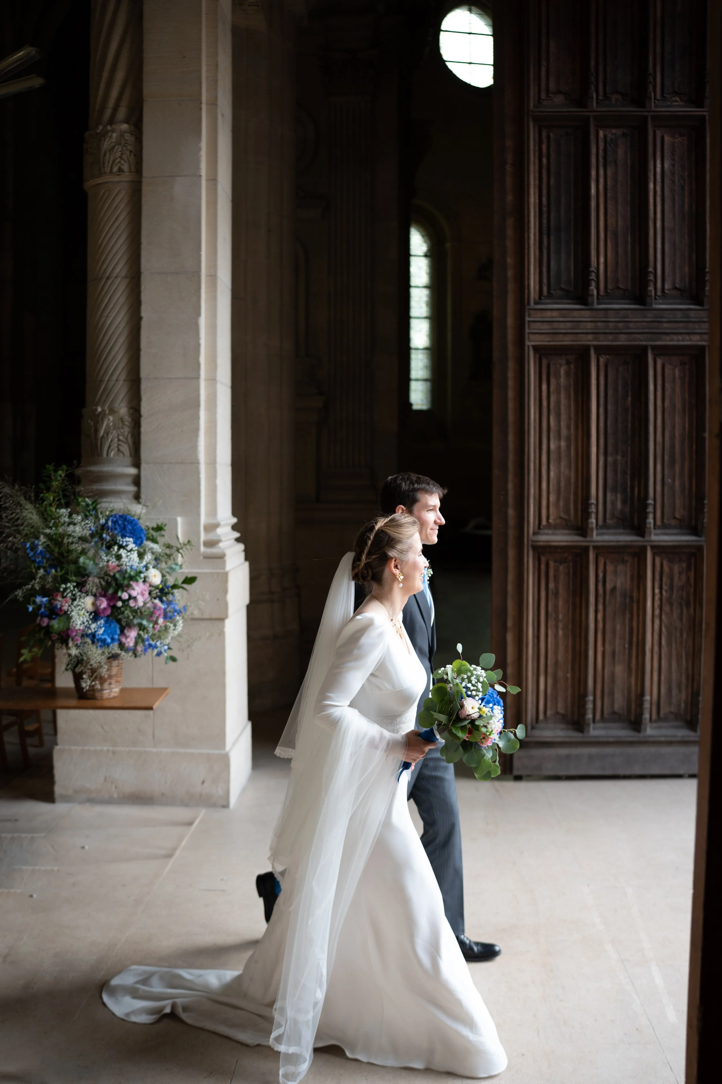 Une jeune femme en robe de mariée et un homme en costume marchent dans une église avec des portes en bois massif, une grande fenêtre et un arrangement floral coloré.