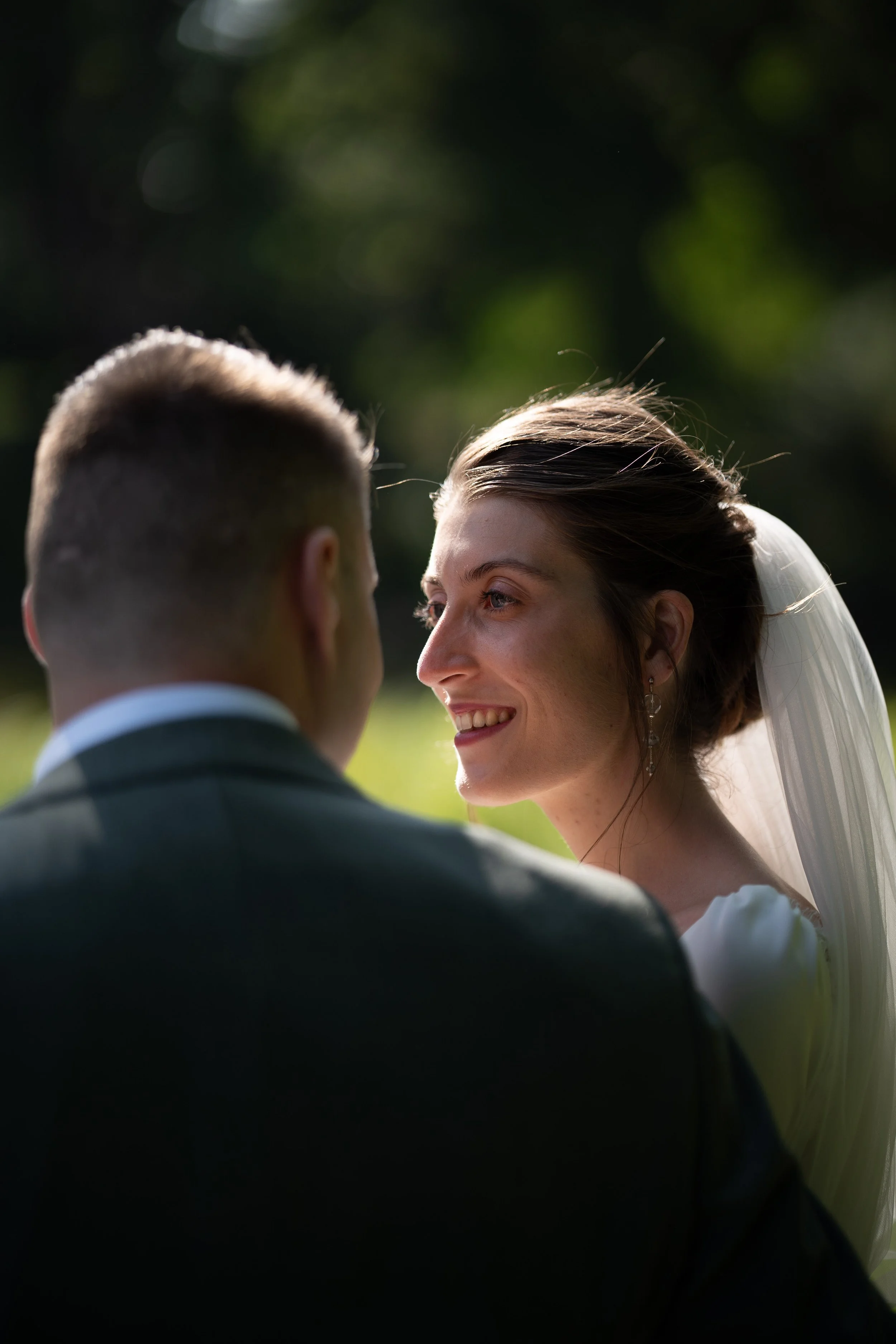 Un couple lors de leur mariage, souriant et se regardant tendrement, en dehors, avec un fond de nature flou.