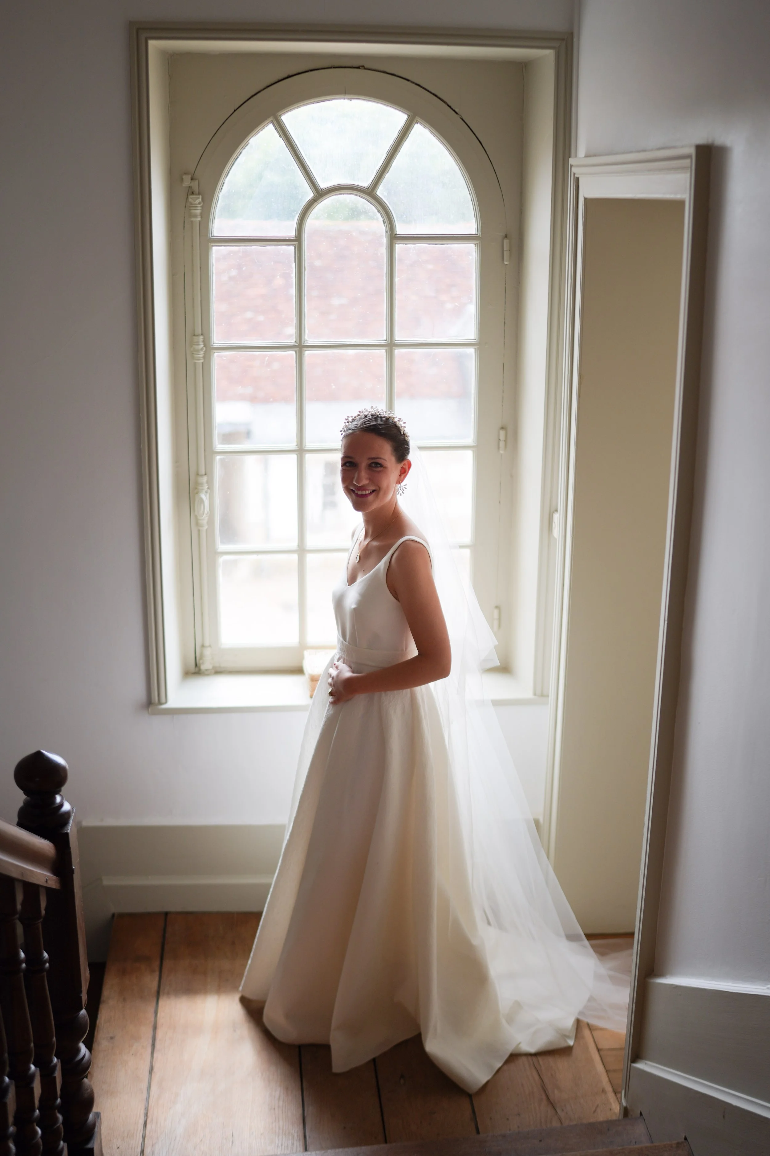Une femme en robe de mariée souriante, debout près d'une grande fenêtre avec de la lumière naturelle, dans une pièce aux murs blancs.