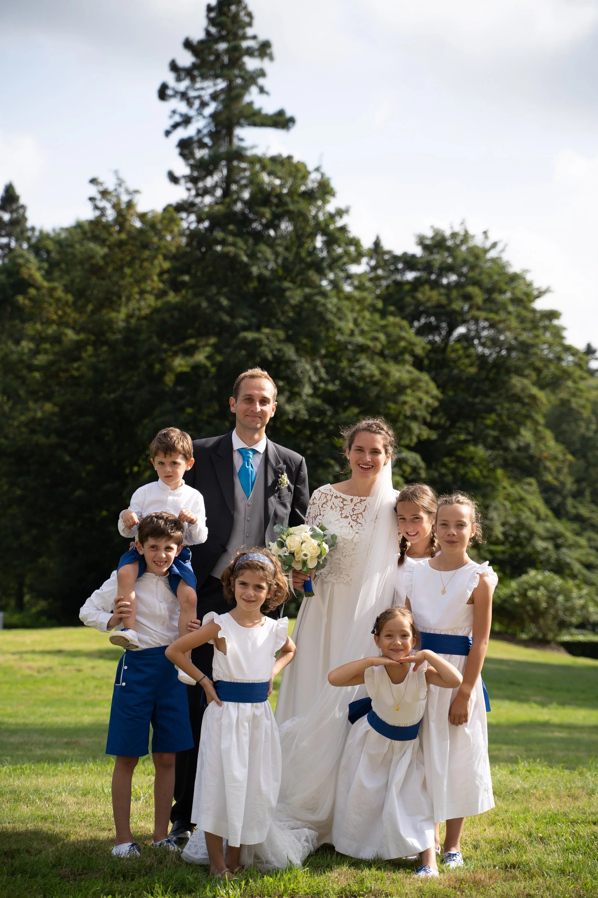 Groupe de personnes lors d'un mariage en plein air, comprenant un couple marié et plusieurs enfants habillés en blanc, posant sur une pelouse avec des arbres en arrière-plan.