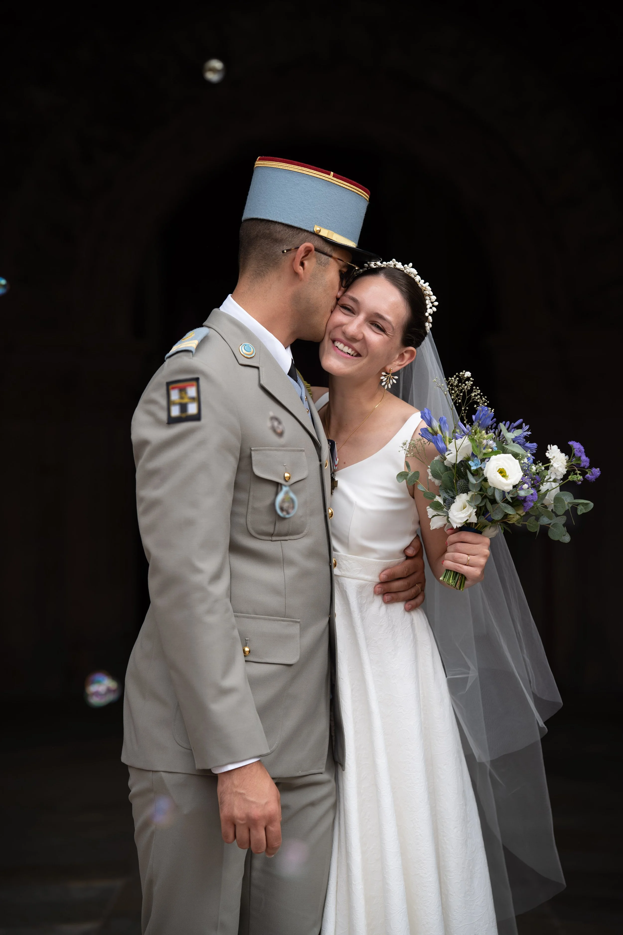 Un couple de mariés, un homme en uniforme militaire et une femme en robe de mariée, s'embrassent tendrement, la femme tenant un bouquet de fleurs. La scène est prise devant un fond sombre.