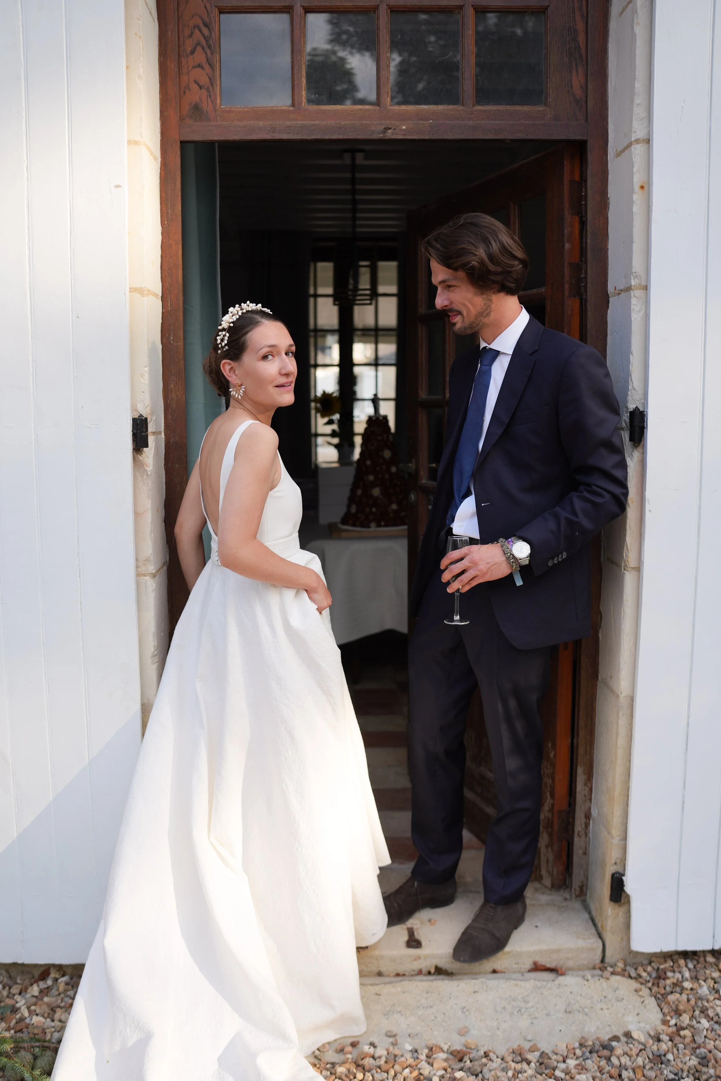 Un couple en tenue de mariage se tient à l'entrée d'une maison en bois, l'homme en costume noir et la femme en robe blanche avec un bandeau de perles sur la tête.