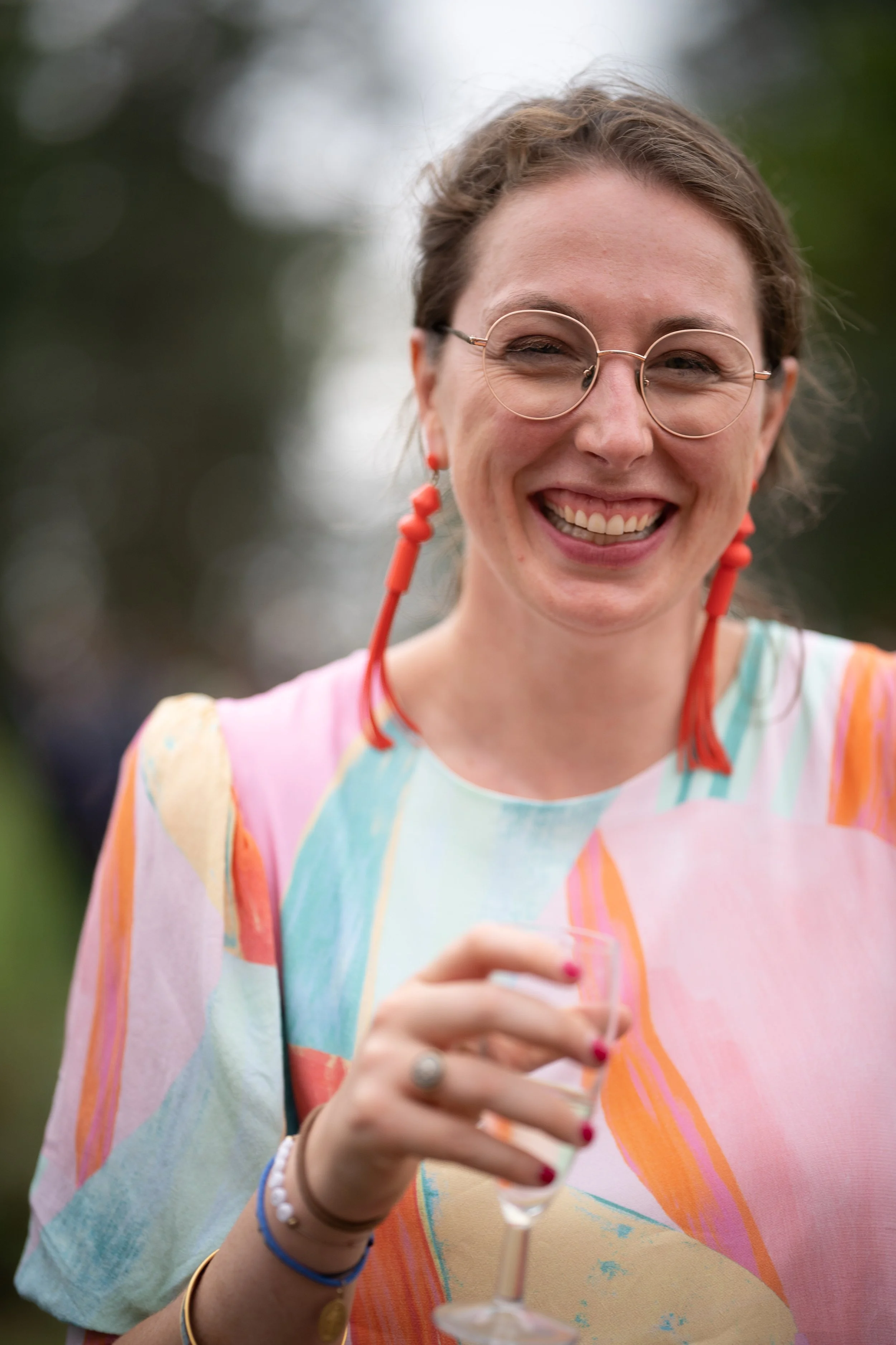 Une femme souriante portant des lunettes rondes, des boucles d'oreilles orange, et un haut coloré en pastel, tenant un verre à vin lors d'une soirée ou d'un rassemblement en plein air.
