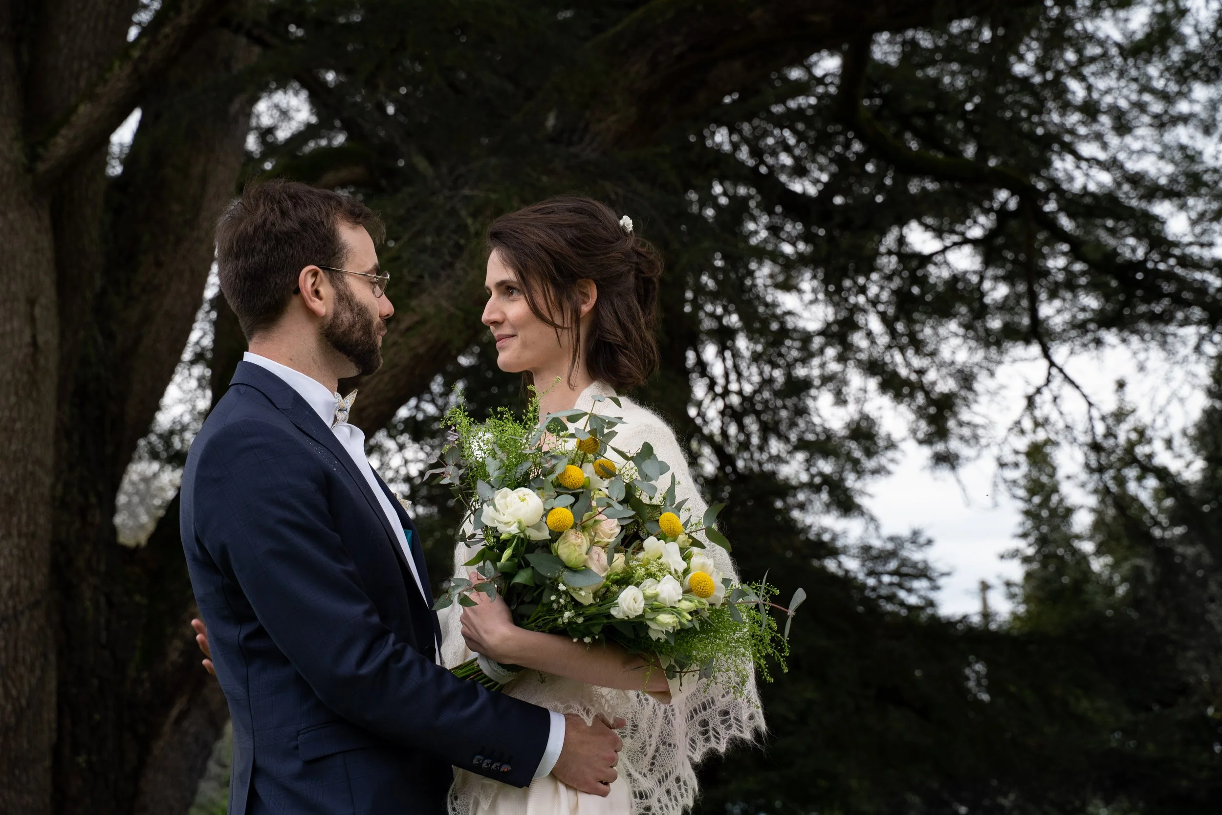 Un couple en tenue de mariage, un homme en costume et une femme en robe de mariée, se regardent avec tendresse sous un arbre. La femme tient un bouquet de fleurs.