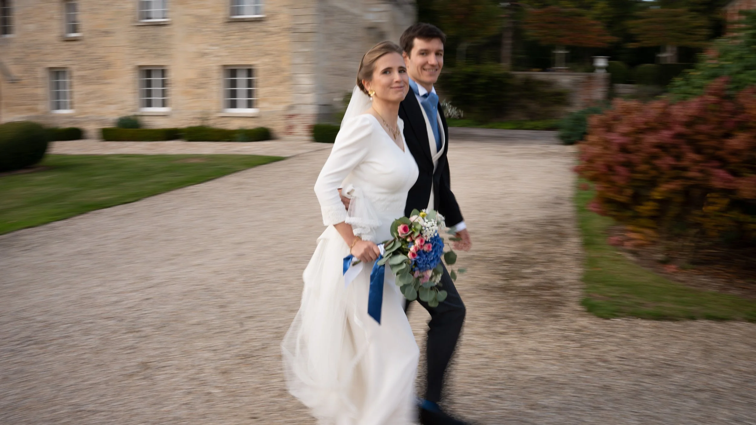 Un couple de mariés marchant main dans la main à l'extérieur, la femme porte une robe de mariée blanche avec un bouquet de fleurs colorées, l'homme porte un costume noir avec une cravate bleue, dans un endroit en plein air avec un bâtiment en pierre 
