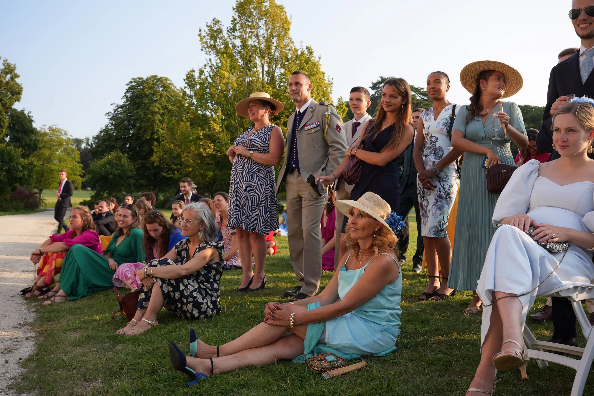 Un groupe de personnes assises et debout dans un parc en pleine journée, portant des vêtements colorés et formels, lors d'un événement en plein air.